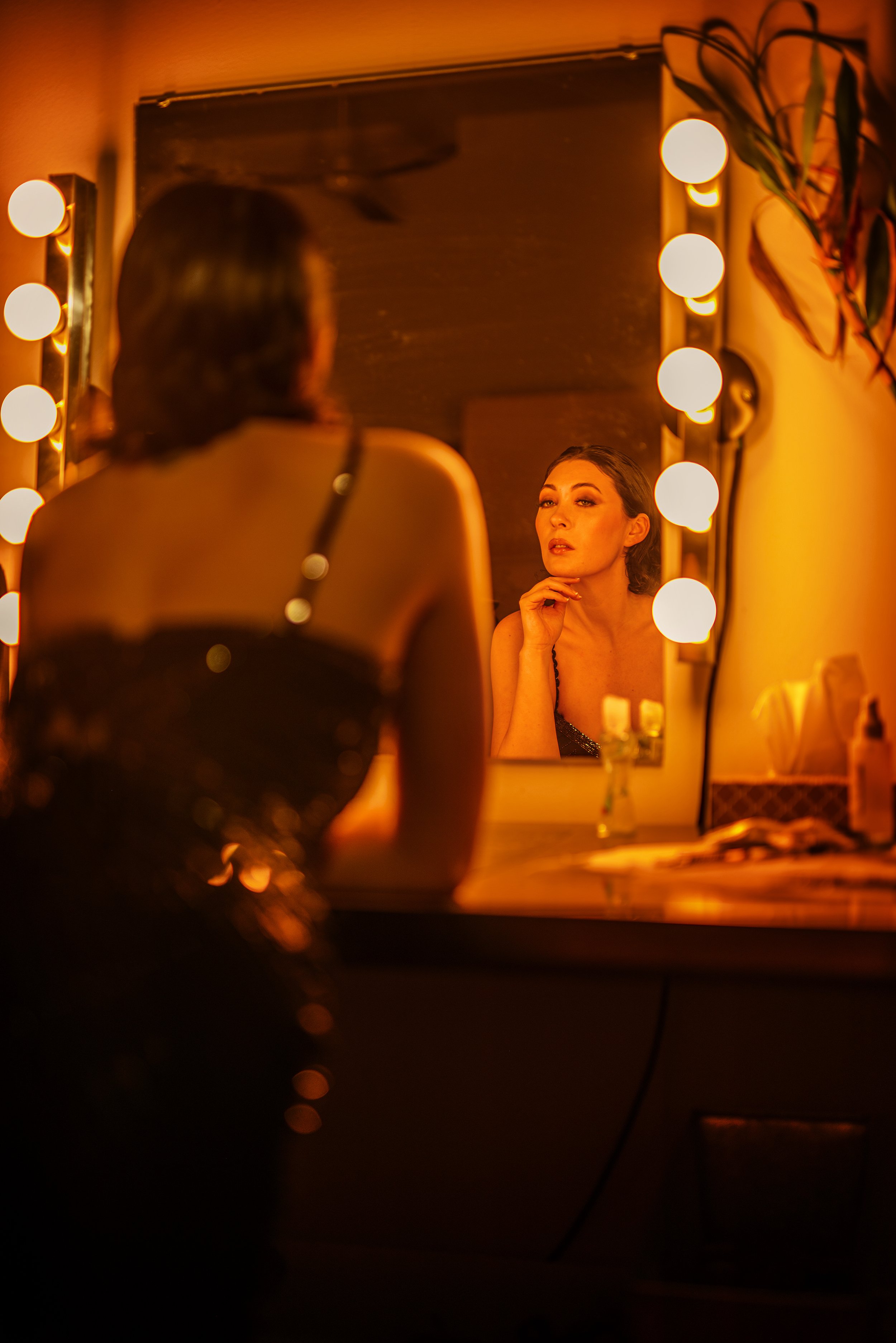 A woman with dark hair looking at her reflection in a mirror with warm, orange lighting. The mirror is surrounded by round light bulbs, and there are some plants and objects on the vanity table.