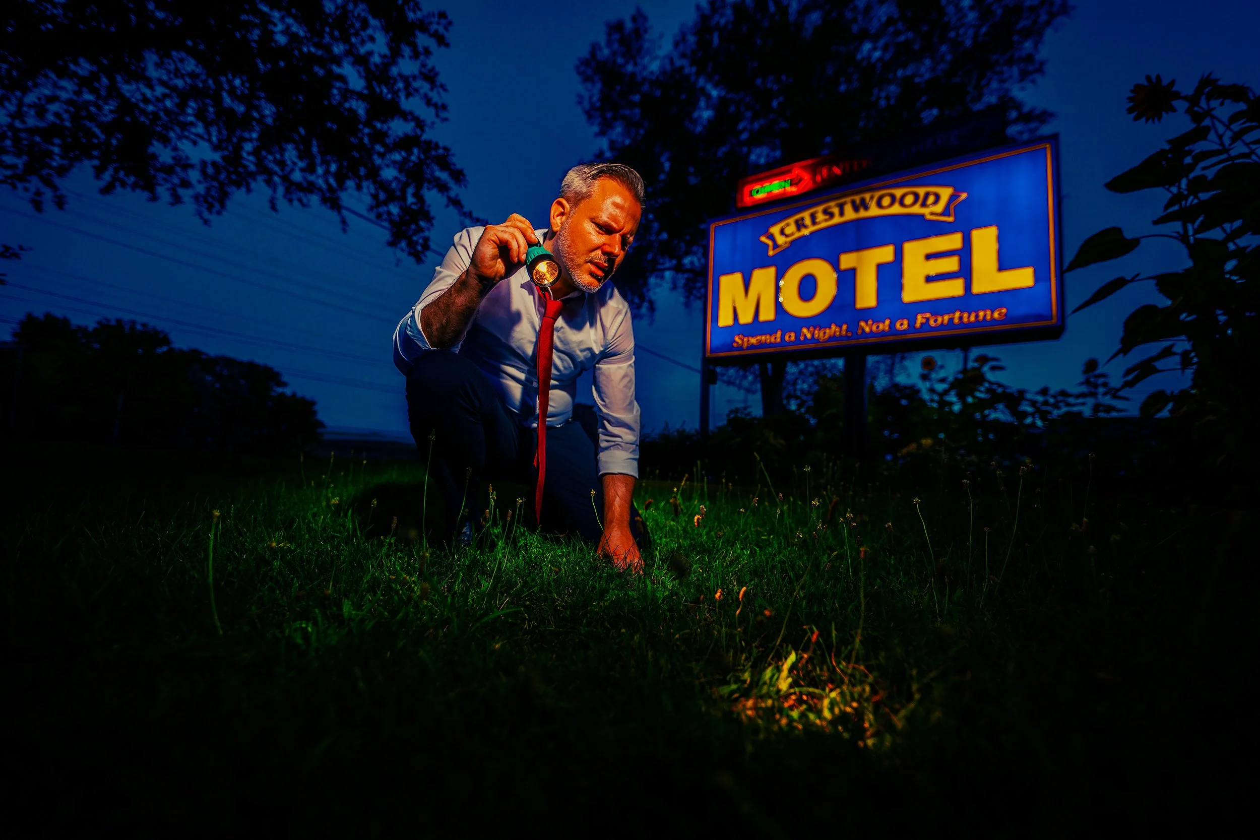 A man in a white shirt and red tie kneels on the grass at dusk, holding a flashlight in his hand, with a motel sign illuminated in the background.