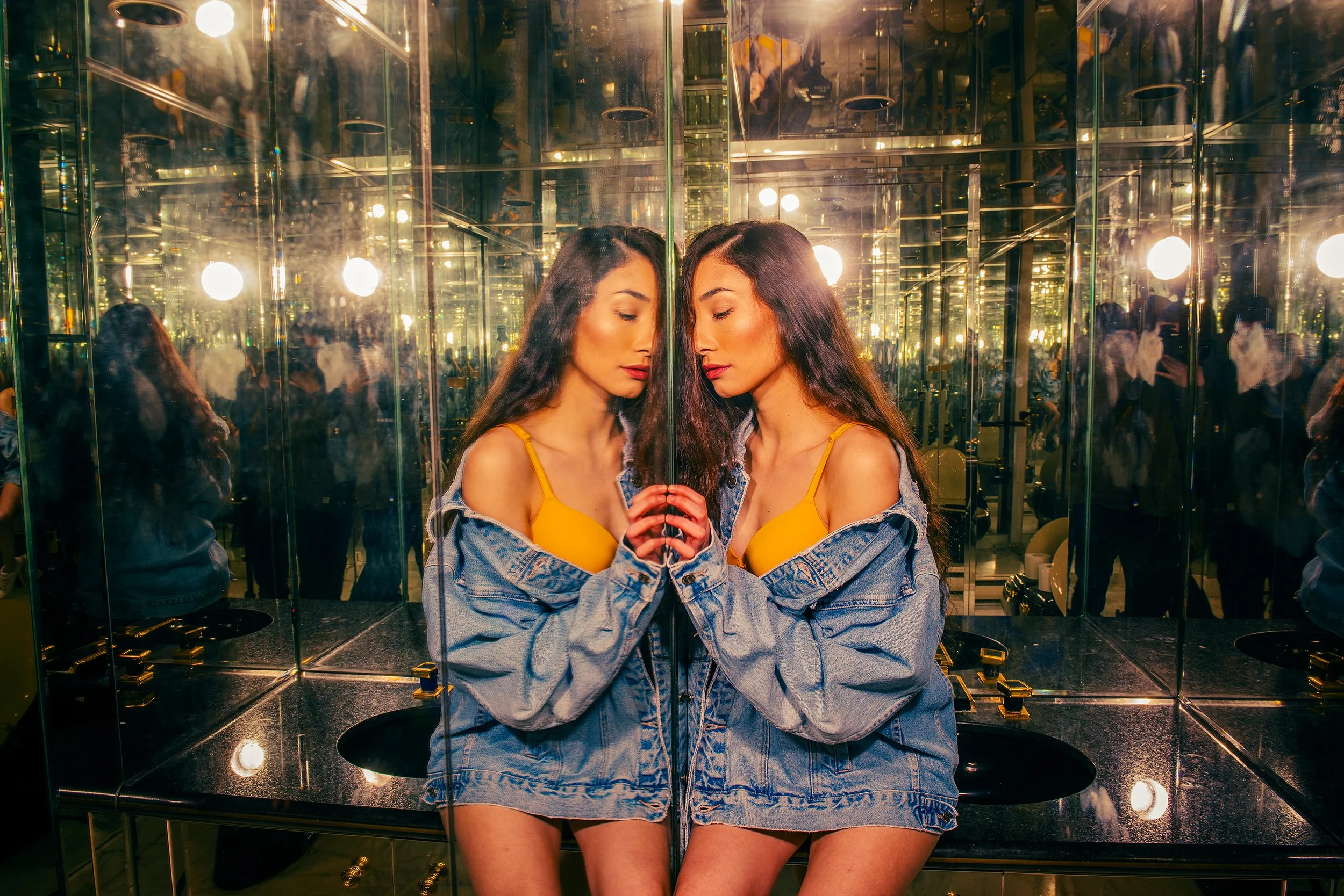 Two young women with long dark hair and casual clothing are standing close to each other, touching foreheads, inside a small, mirrored room with multiple lights and glass walls. Their eyes are closed, and their hands are touching.