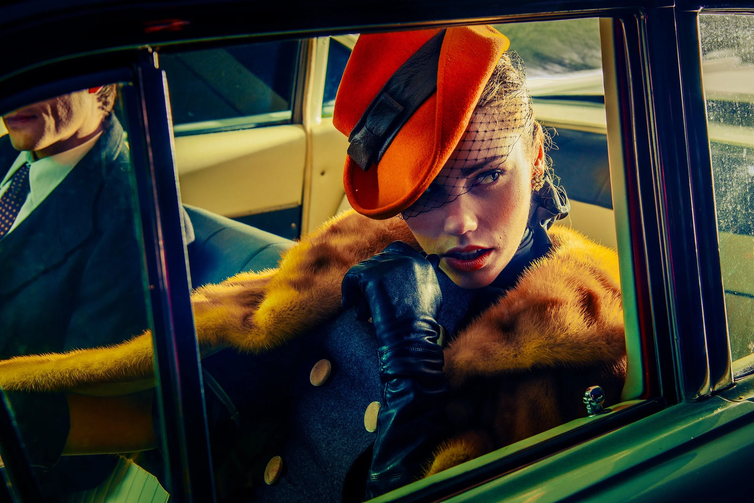 A woman wearing a vibrant orange hat with black accents, a bright orange faux fur coat, and black gloves sitting in the front seat of a vintage car, with a young man in a suit and tie seated beside her. The woman's face is partially visible, and she 