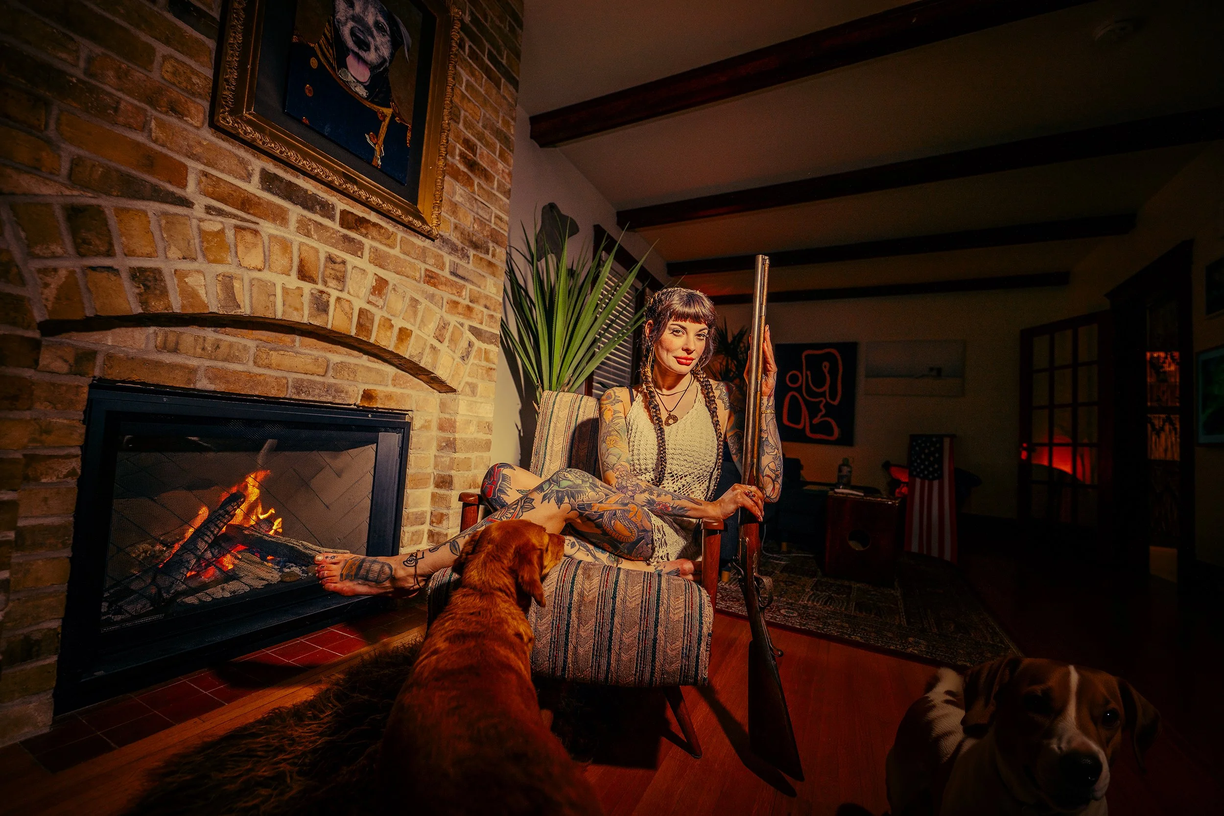A woman with tattoos, dark hair, and bangs sitting by a fireplace holding a long pipe. There are two dogs in the scene, one lying on the woman's lap and the other in the foreground. The background features brick walls, artwork, a tall plant, and warm