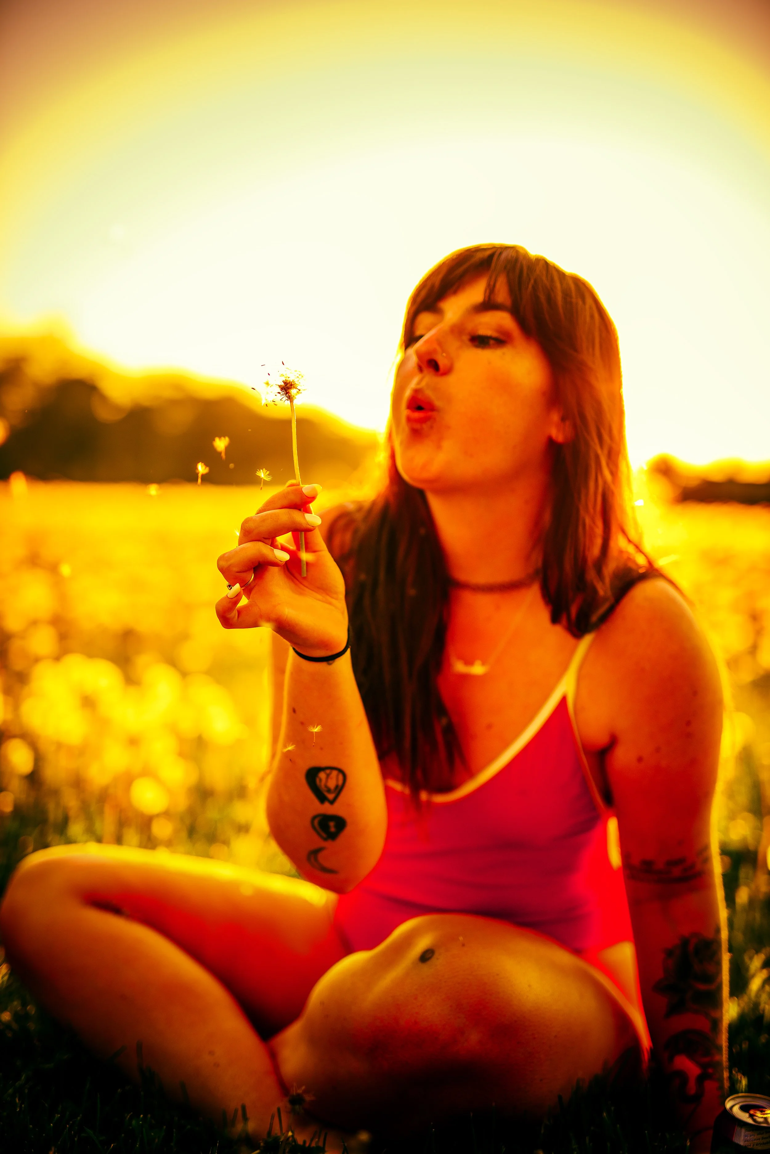 A woman sitting outdoors during sunset, blowing on a dandelion, with tattoos on her arms, surrounded by a field of grass and flowers.