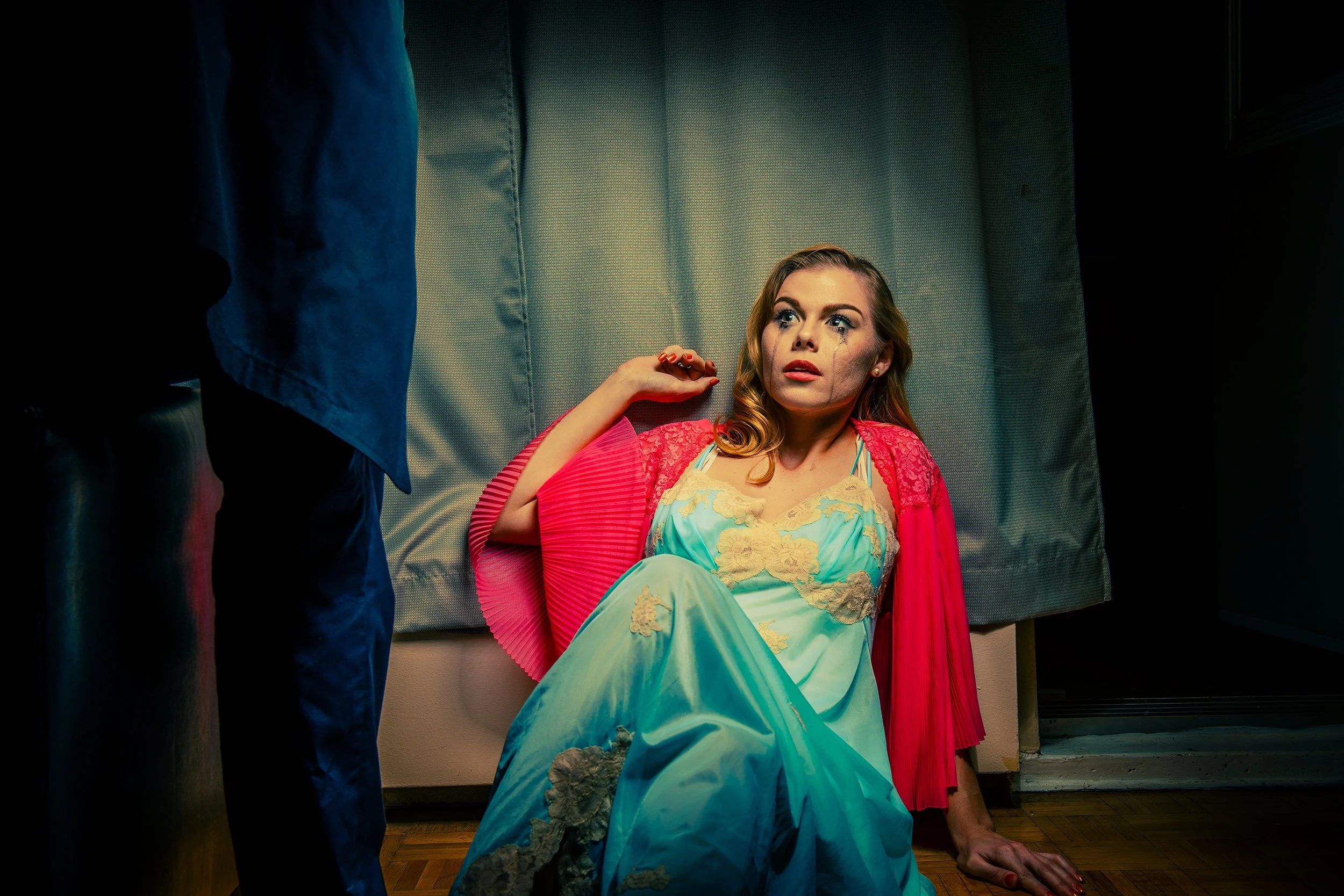 A woman with makeup and tears on her face sitting on the floor against a wall, wearing a colorful gown, with someone standing nearby partly visible.