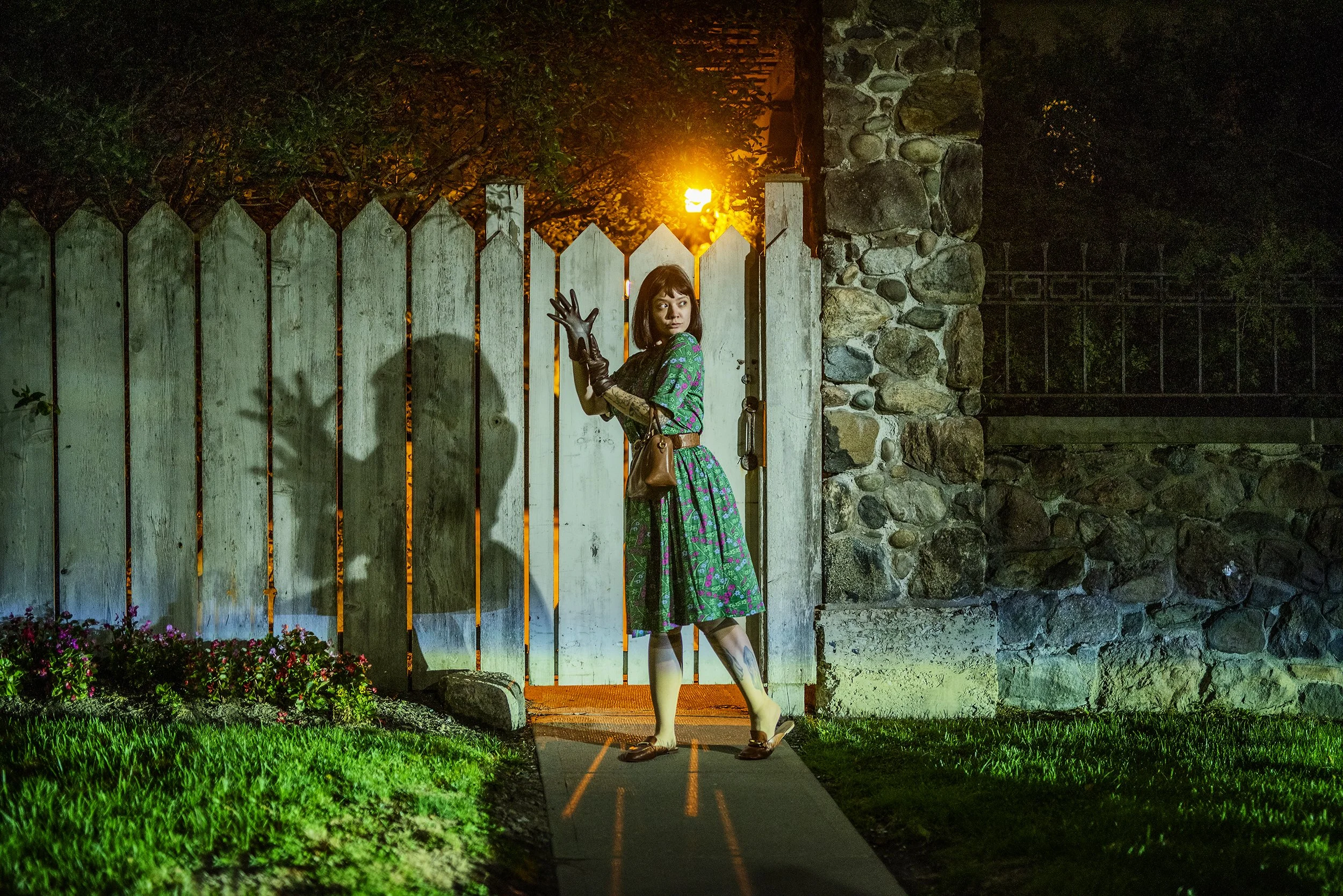 A woman standing at a gate at night, illuminated by a warm light behind her. She is wearing a colorful dress, a brown purse, and gloves, with one hand on the gate and the other raised in a gesture. The background includes a stone wall, greenery, and 