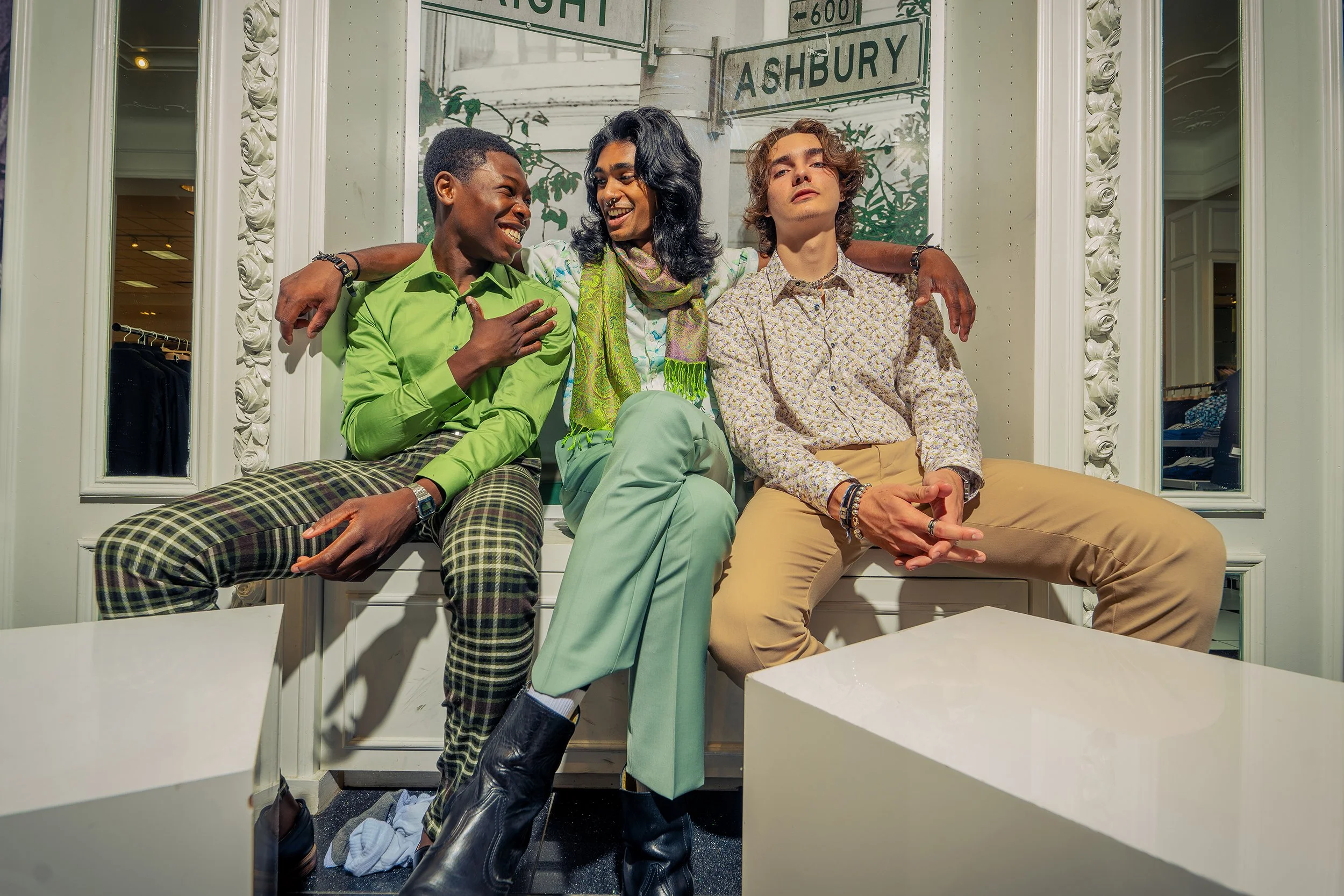 Three young adults sitting on a ledge outside a building, smiling and engaging with each other, with street signs for Ashbury and a high street in the background.