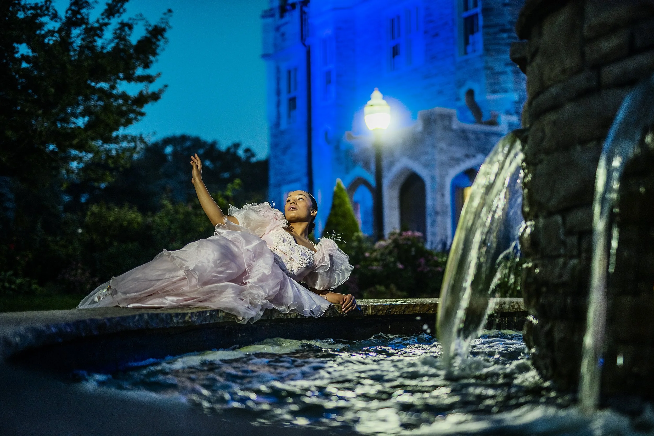 A woman in a pink, ruffled dress lying on a ledge near water, reaching out with her hand, with a castle-like building illuminated in blue in the background at night.