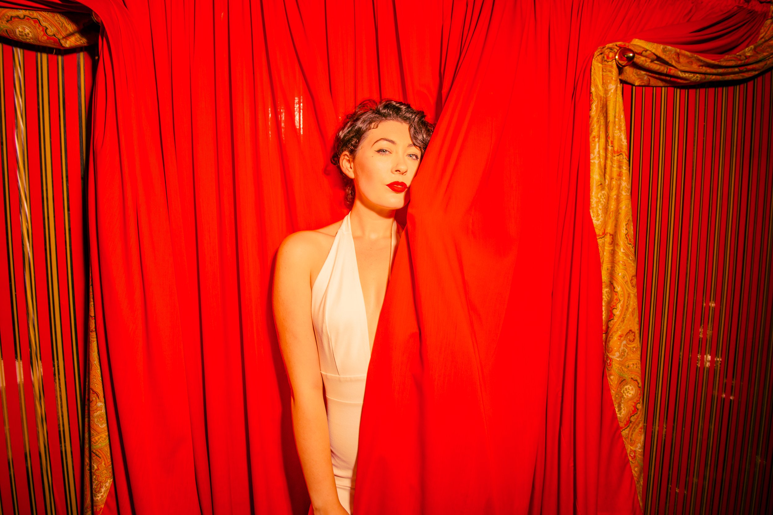 A woman with short curly hair and red lipstick peeks out from behind a red curtain in a vintage-style dressing room with ornate gold and striped fabric on the sides.