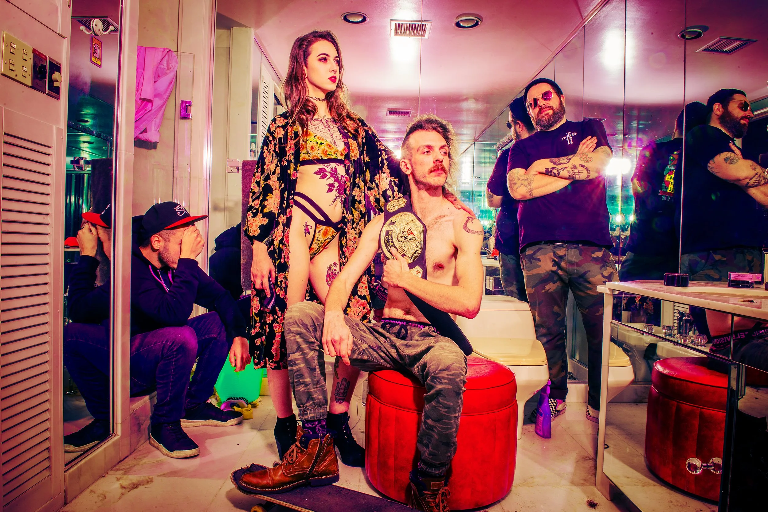 Group of five people in a room with a mirror wall reflection, with two seated and three standing. The seated man has tattoos, a mustache, and is holding a championship belt. The woman standing has long wavy hair and wears lingerie with a floral robe.