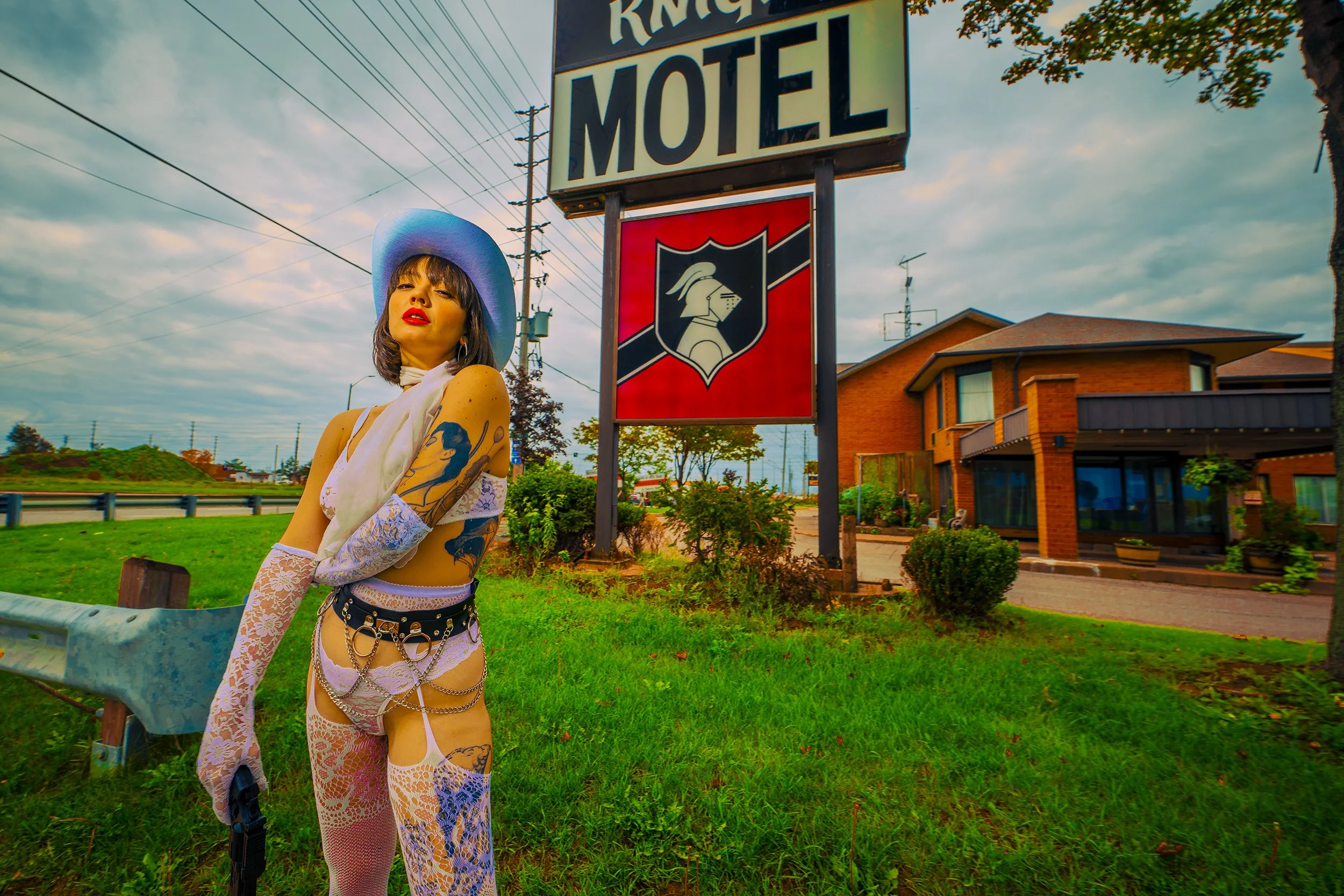 A woman with tattoos wearing a blue wide-brimmed hat, white lace gloves, and fishnet stockings, stands on a grassy roadside near a motel sign with a red shield featuring a knight's helmet logo, under a cloudy sky.