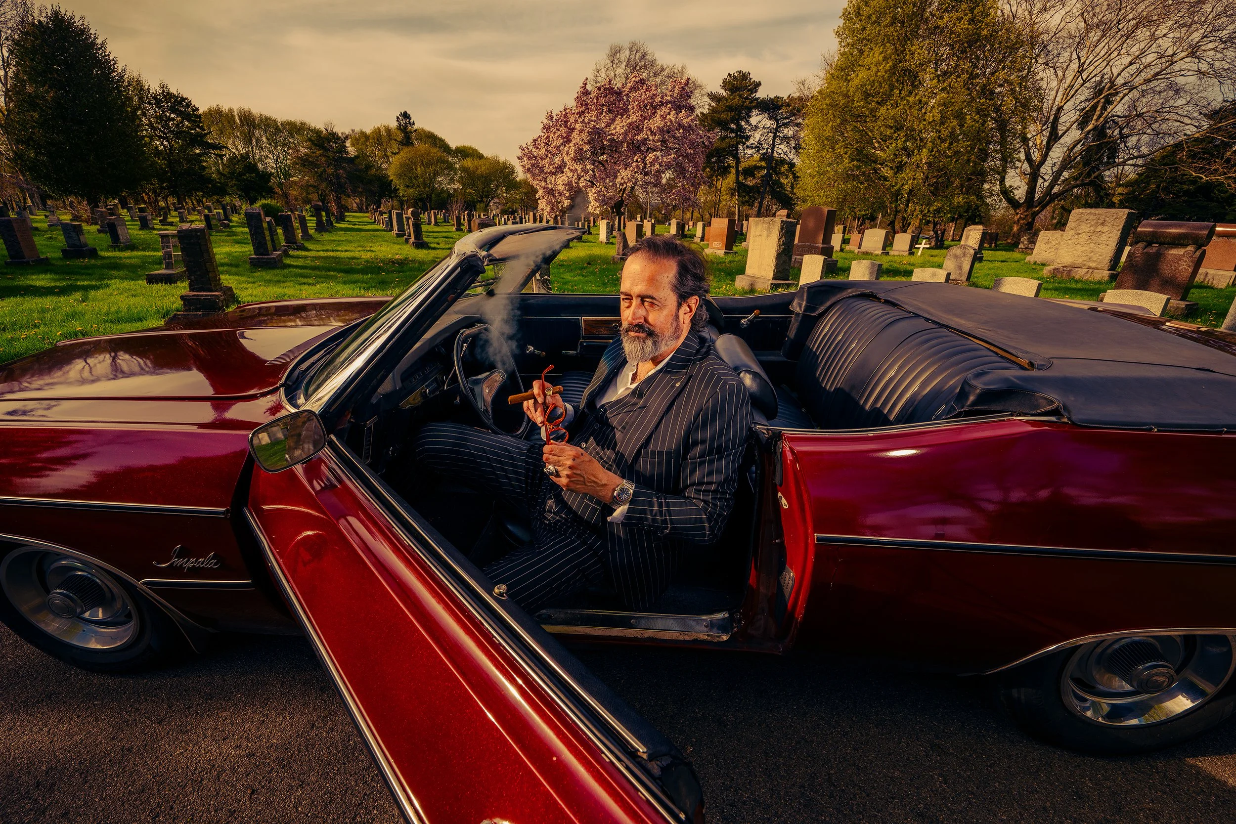 A man with a beard and mustache sitting in the driver's seat of a vintage red convertible car, holding a cigar. The background features a cemetery with numerous gravestones and trees, and a sky with a warm, golden hue.