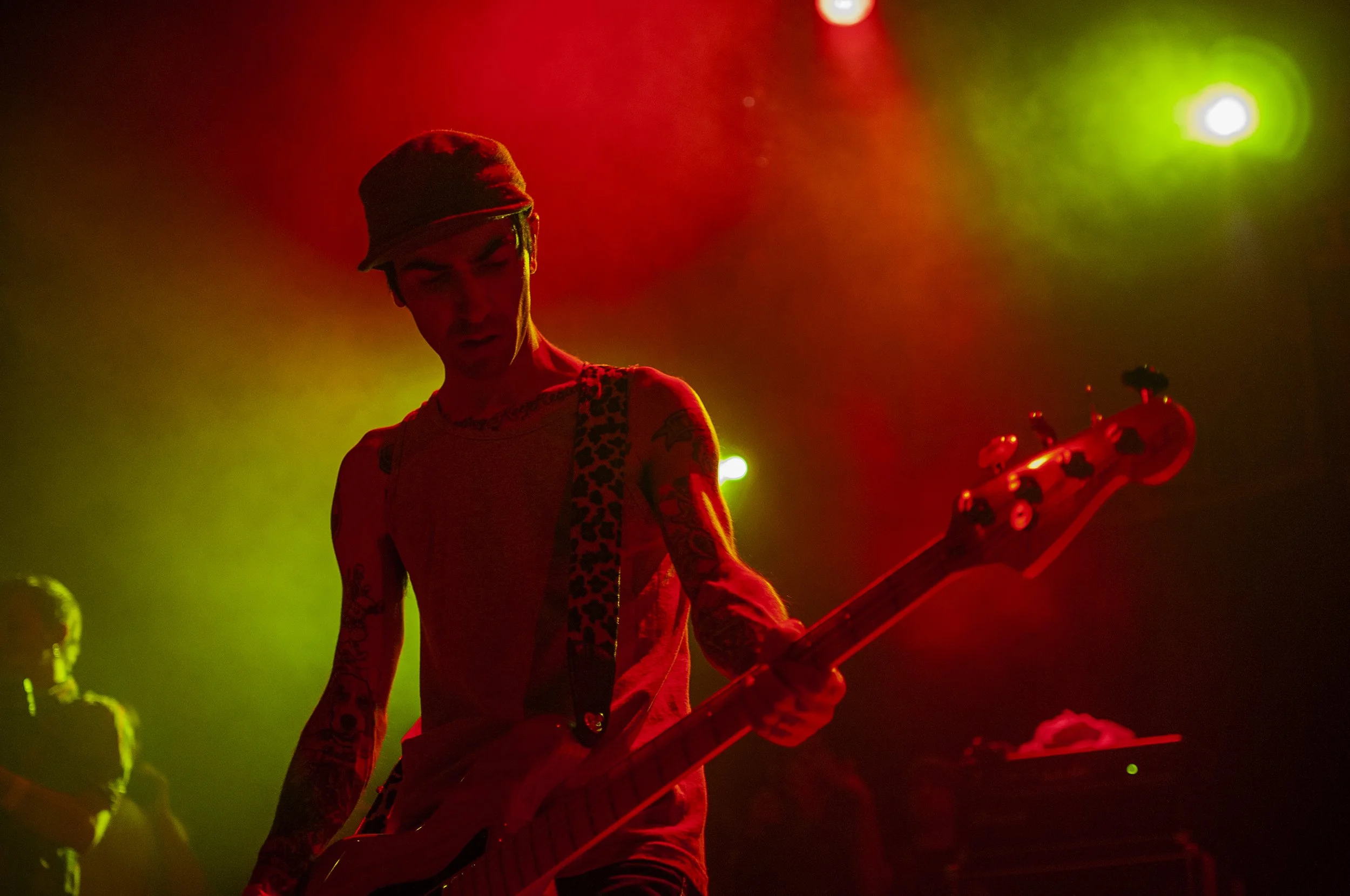 A musician playing a guitar on stage with colorful stage lights, wearing a cap and sleeveless shirt with tattoos visible on their arms.