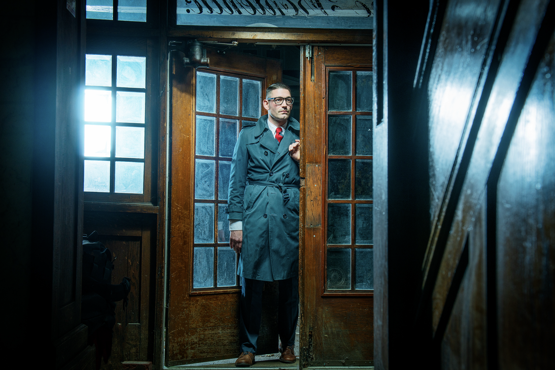 A man in a trench coat and glasses standing partially behind a wooden door with glass panels, in a rustic interior with wooden walls and windows.