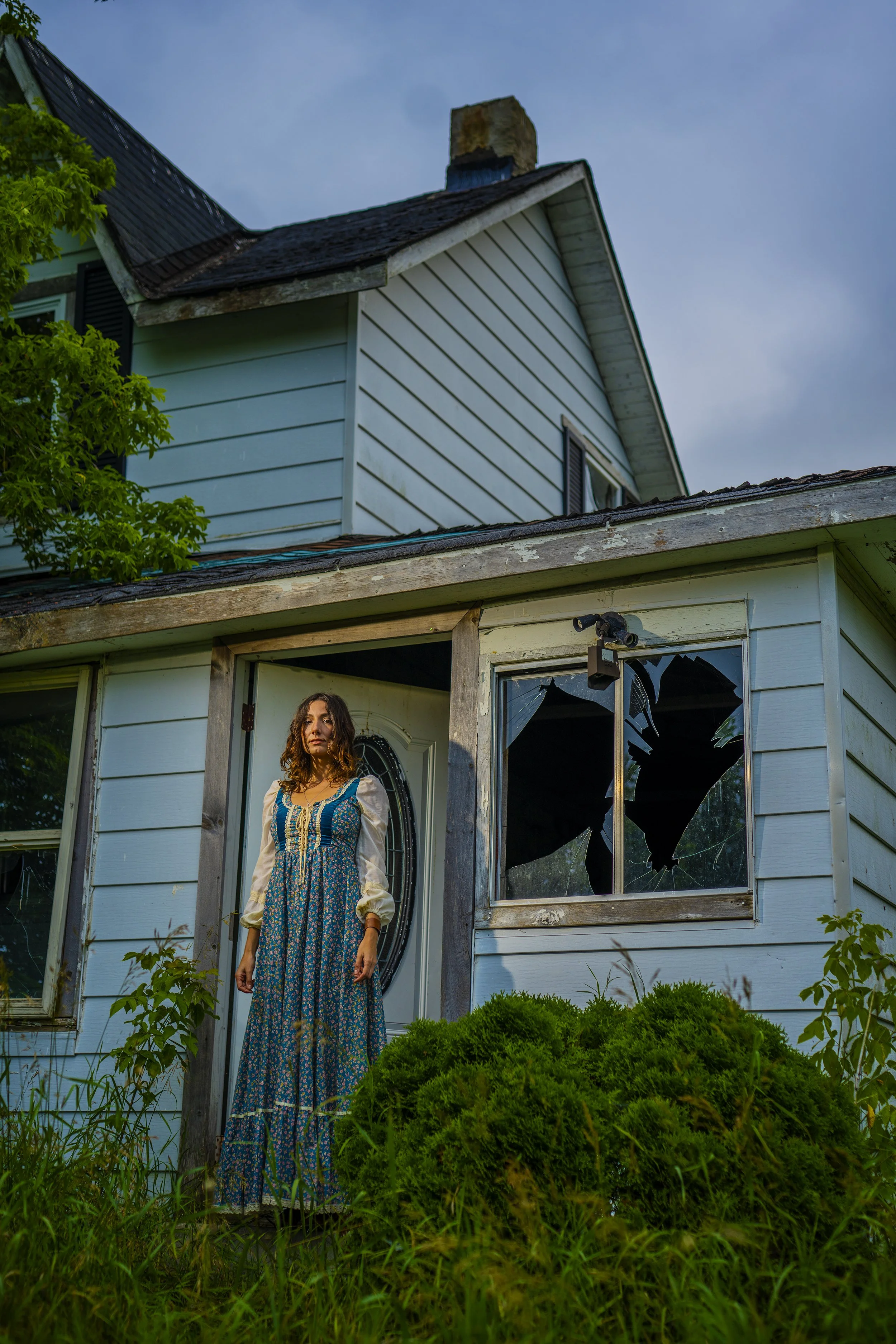 A woman with long curly hair dressed in a long blue floral dress stands in front of a dilapidated house with broken windows. The house has peeling paint and the roof appears to be damaged. The scene is outdoors with green bushes and grass in the fore