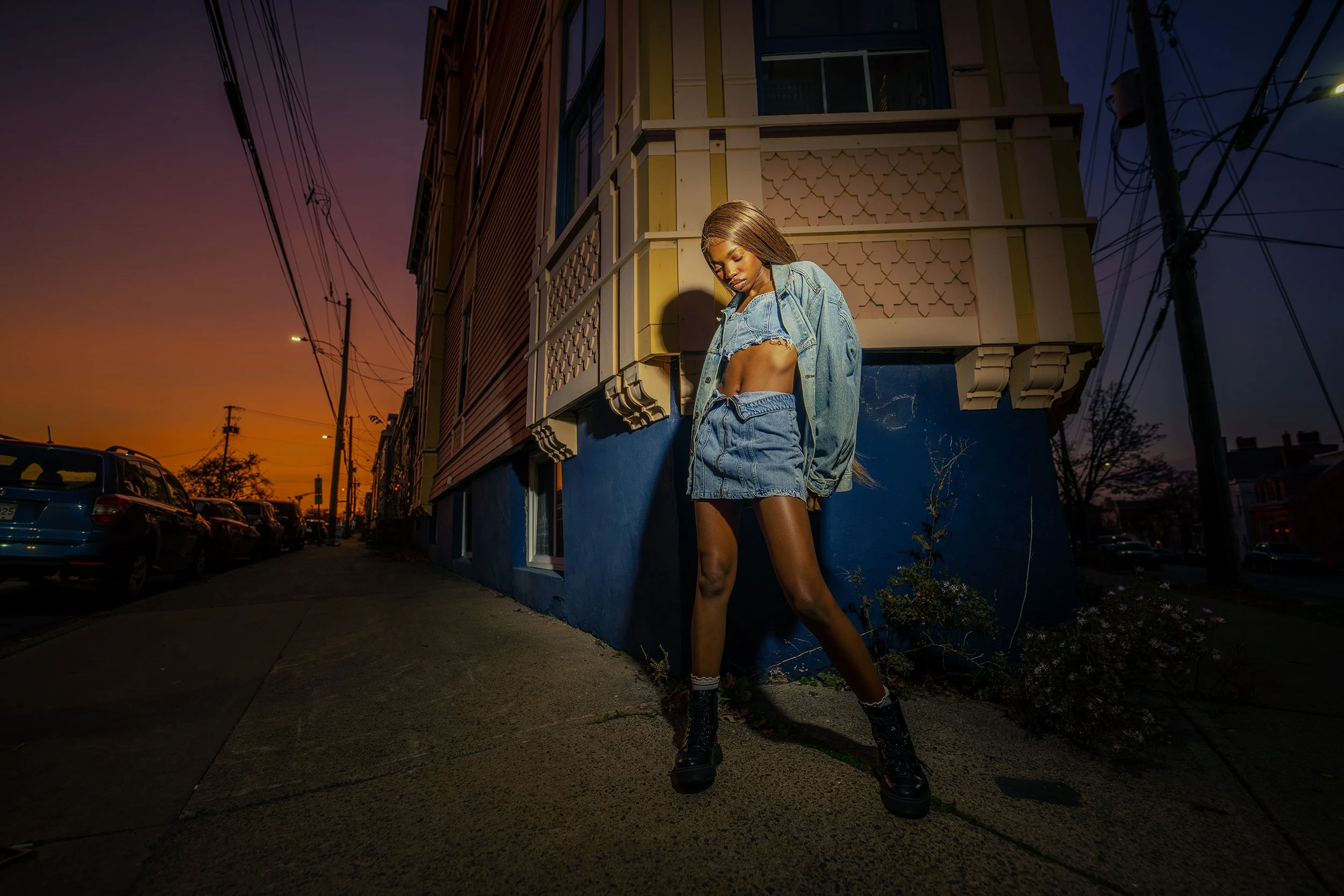 A young woman with long hair wearing a cropped denim jacket and matching denim skirt, standing on a sidewalk at sunset near a colorful building and parked cars.