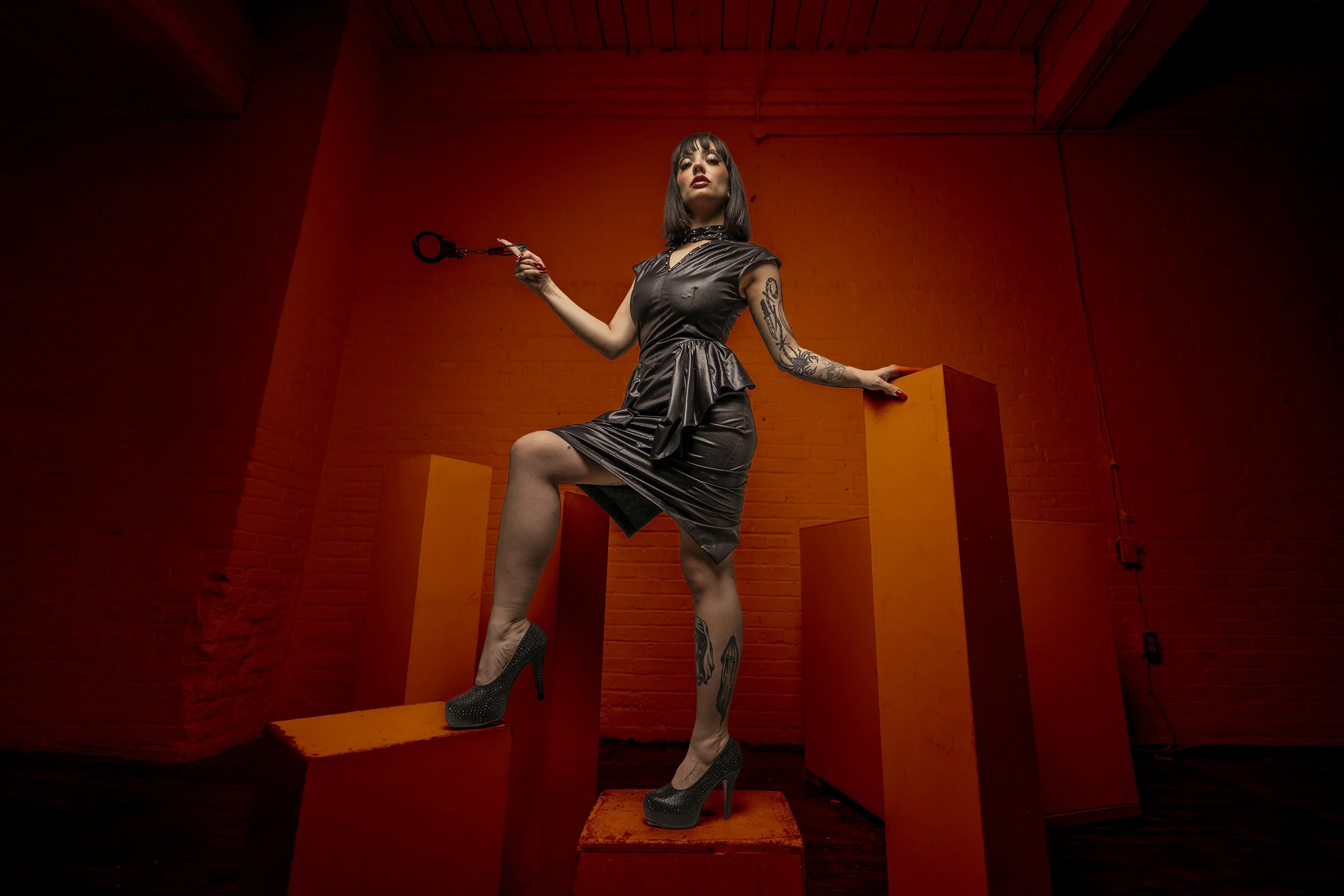 A woman with dark hair and tattoos wearing a black dress and high heels, holding handcuffs, standing on orange blocks in a red-lit room.