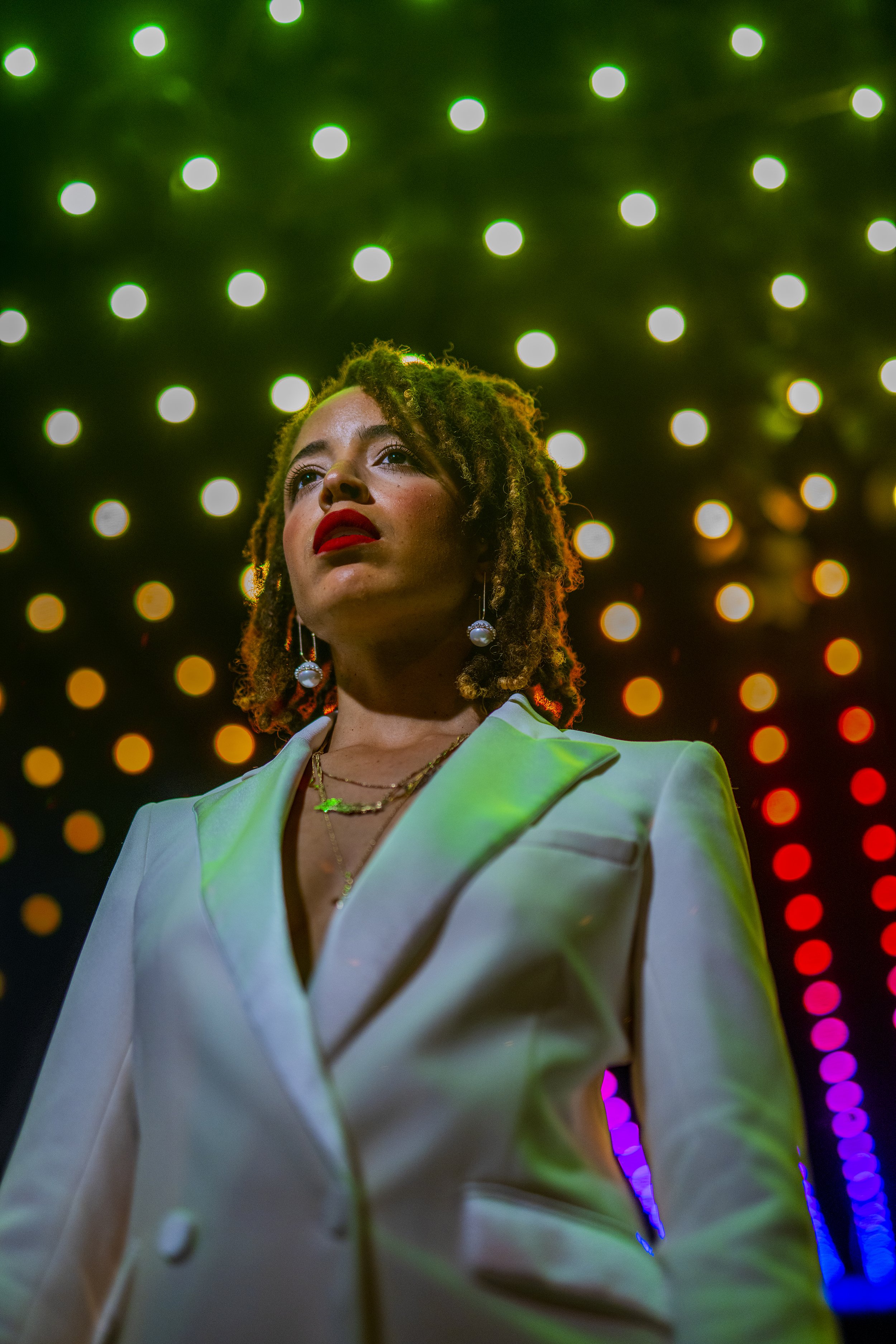 A woman with curly dreadlocks wearing red lipstick and pearl earrings, standing in front of a background of colorful, blurred circular lights.