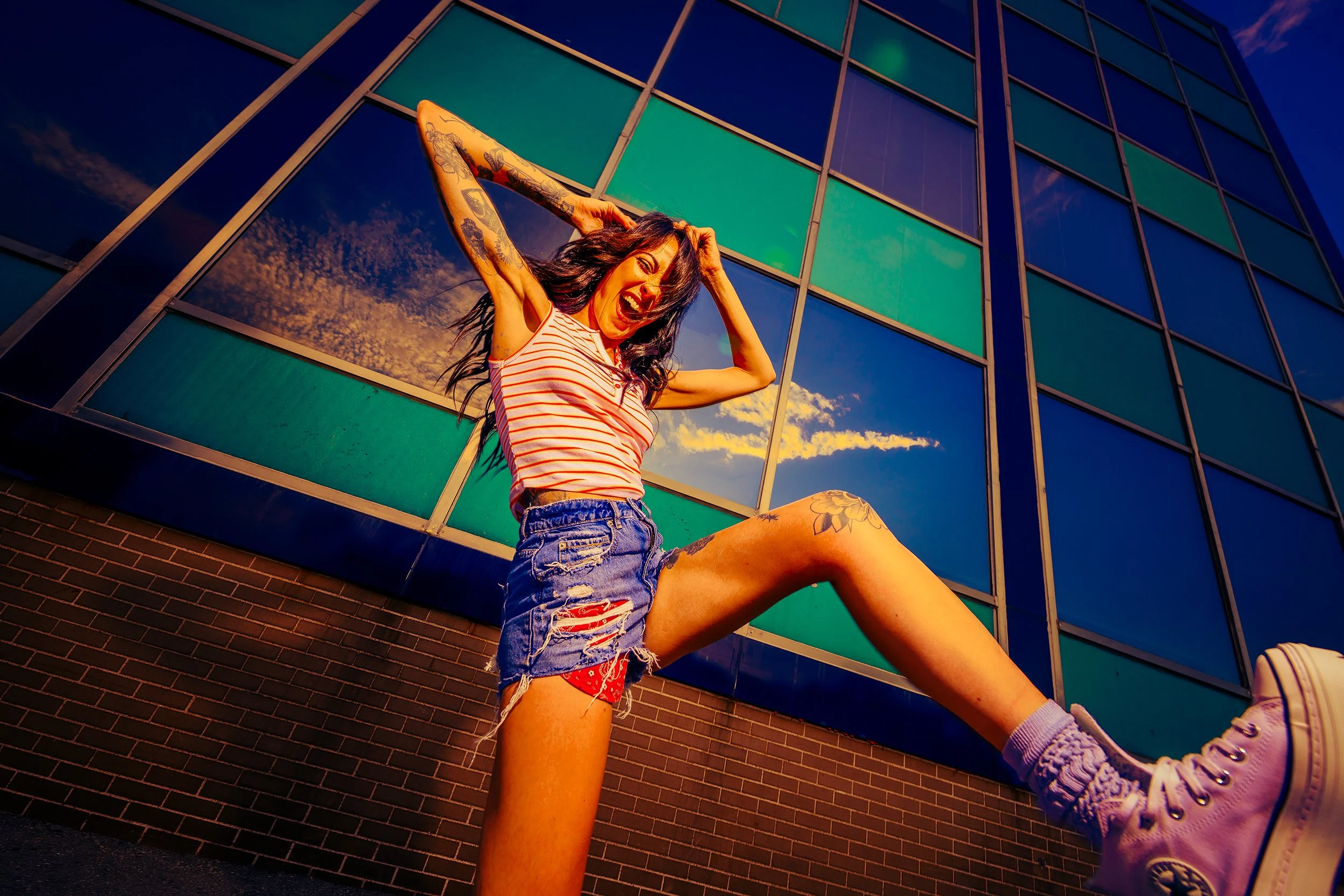 A young woman in a striped tank top and ripped denim shorts smiling and stretching against a reflective glass building during sunset.