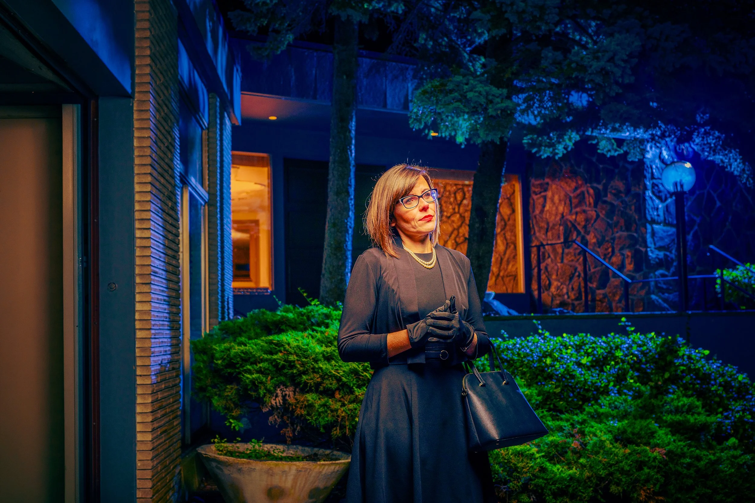 A woman dressed in a black dress with gloves and glasses stands outside at night, holding a black purse, with colorful exterior lighting illuminating her and the plants around her.