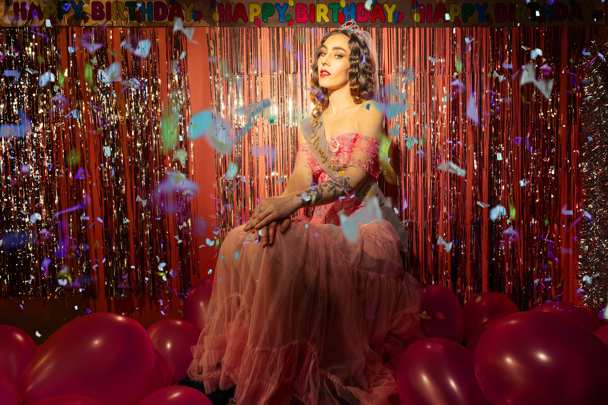 A woman in a pink dress and tiara sitting among red balloons at a birthday celebration with colorful decorations, including a 'Happy Birthday' banner and shiny streamers, with confetti falling around her.