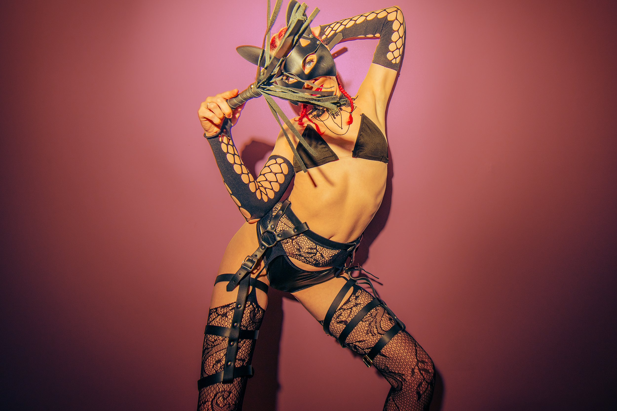 Person in black lingerie, fishnet stockings, and harnesses wearing a horse mask, holding a crop, against a pink background.