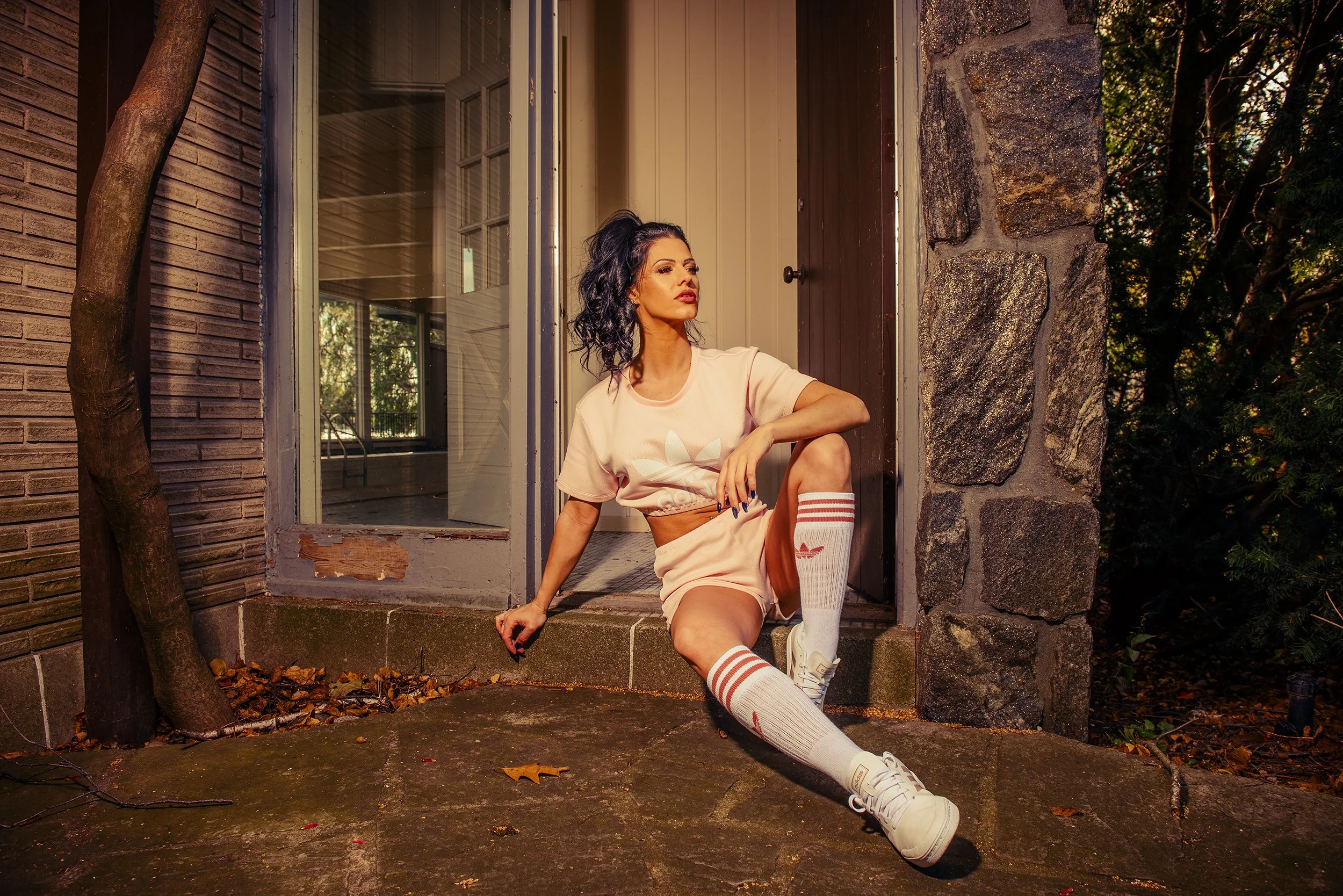 A woman with black curly hair sitting on a porch step, wearing a beige T-shirt, beige shorts, and white socks with red stripes, in front of a house with a stone and brick wall, surrounded by trees and autumn leaves.