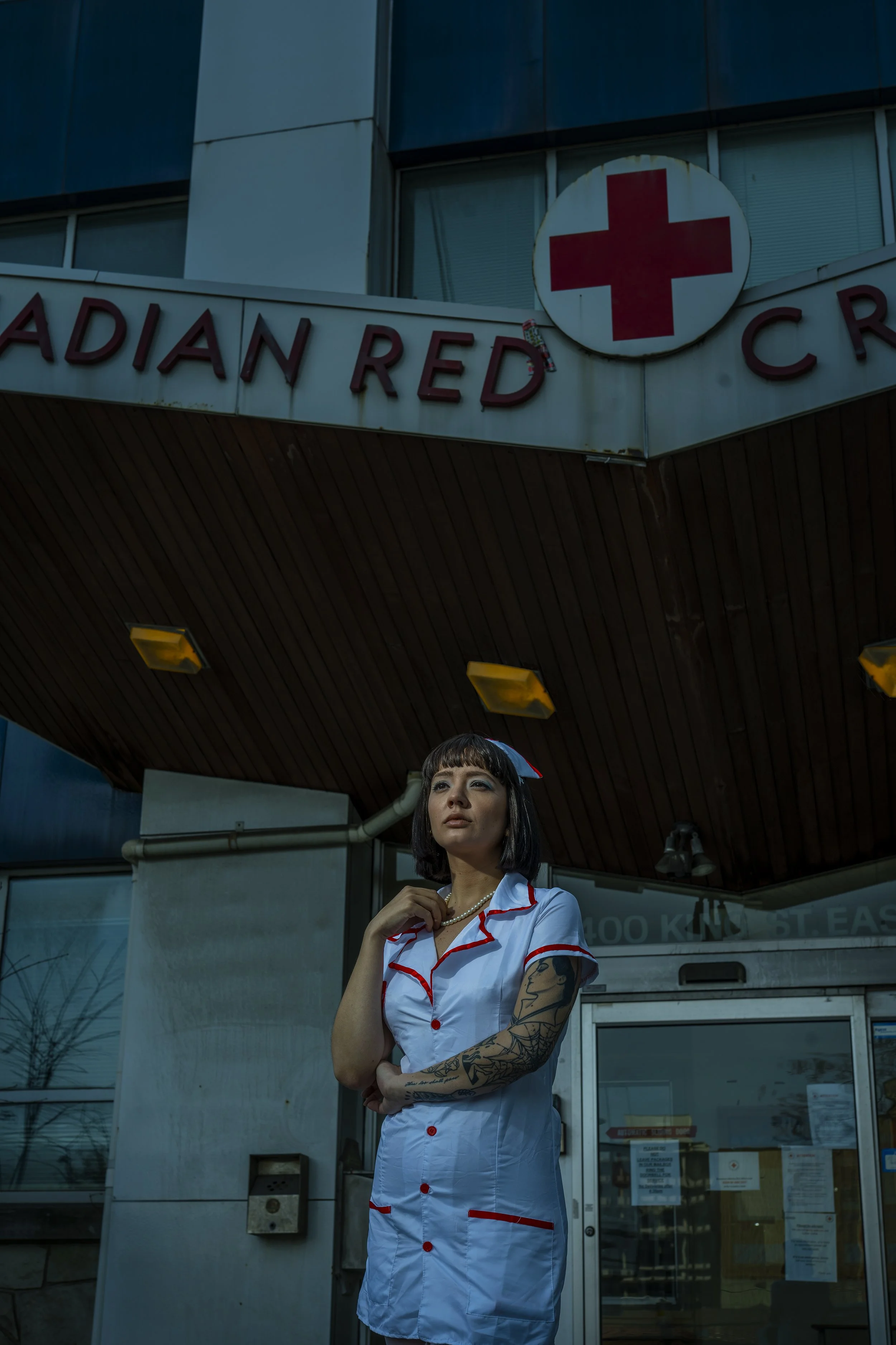 A woman dressed as a nurse with a tattoo on her arm, standing outside a building with a sign showing a red cross and the words 'Adian Red' partially visible. She is wearing a white dress with red trim and has short black hair.