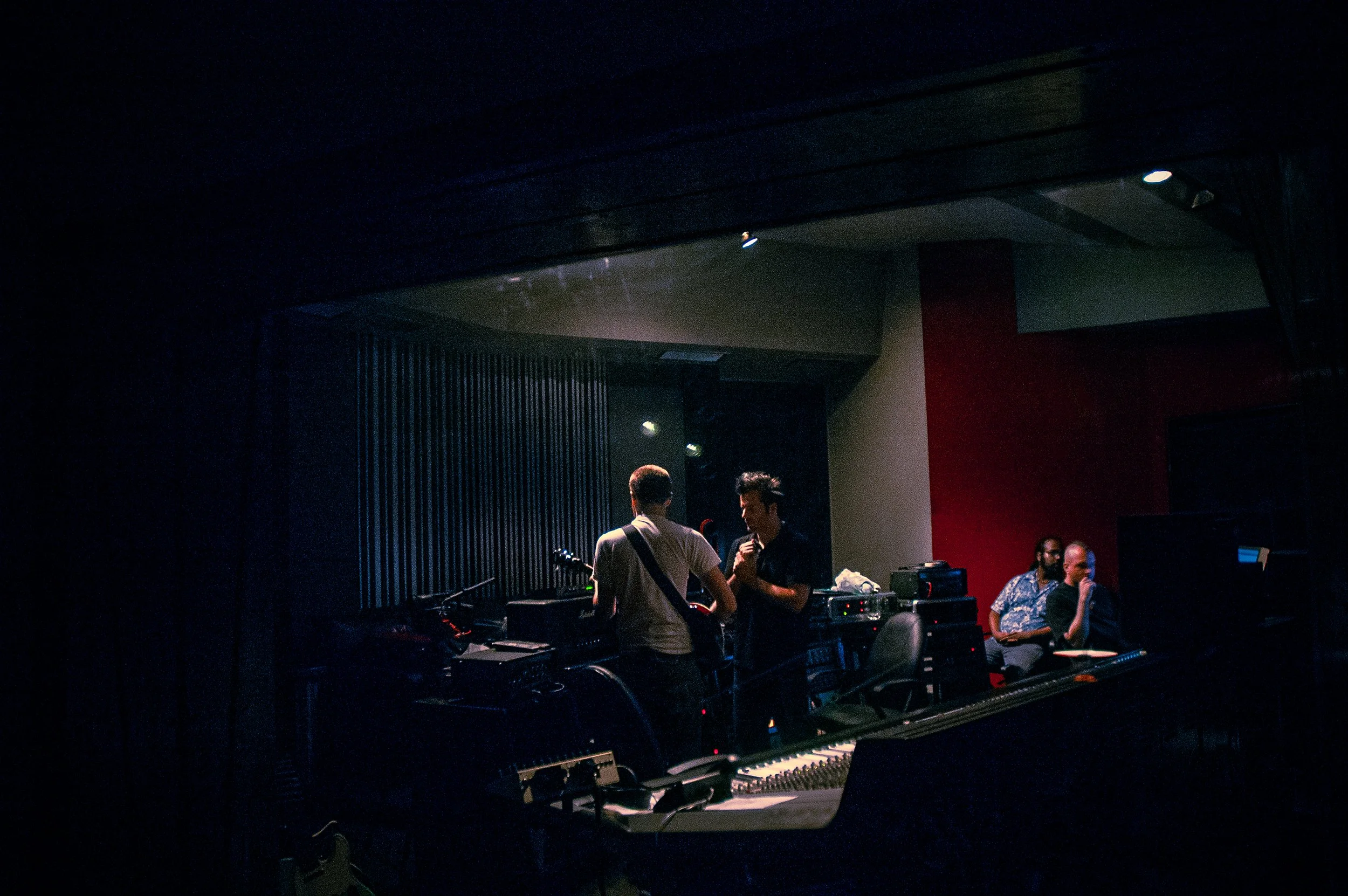 People gathered in a dimly lit room with a piano in the foreground, some individuals appear to be engaged in conversation near audio equipment, with a dark ceiling and walls.