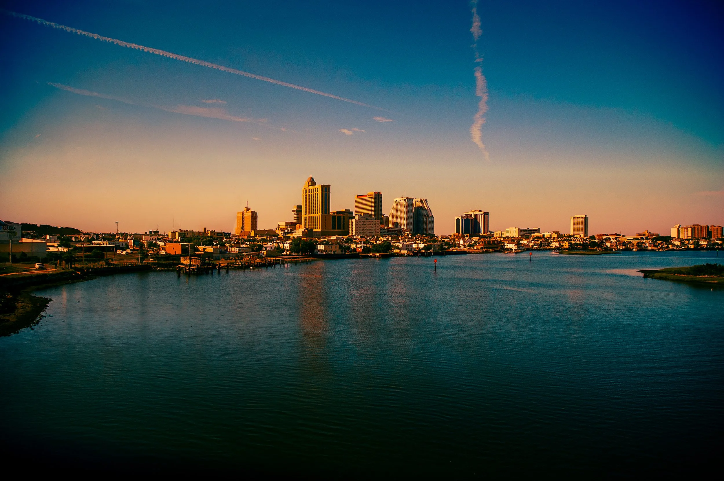 City skyline during sunset with high-rise buildings, water in foreground, and contrails in the sky.