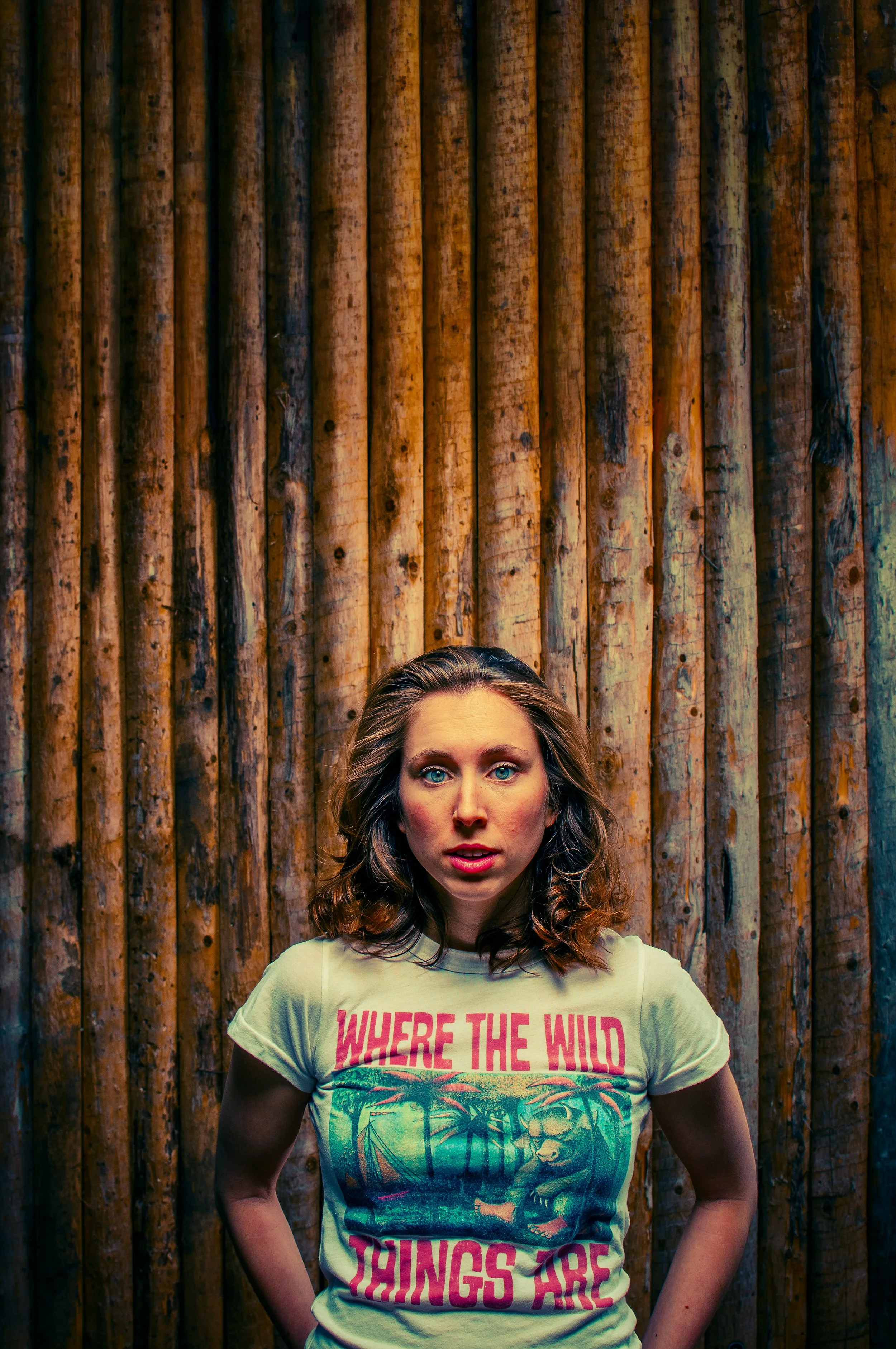 A woman with wavy brown hair and blue eyes wearing a yellow T-shirt with pink text that says 'WHERE THE WILD THINGS ARE' standing in front of a wooden fence.