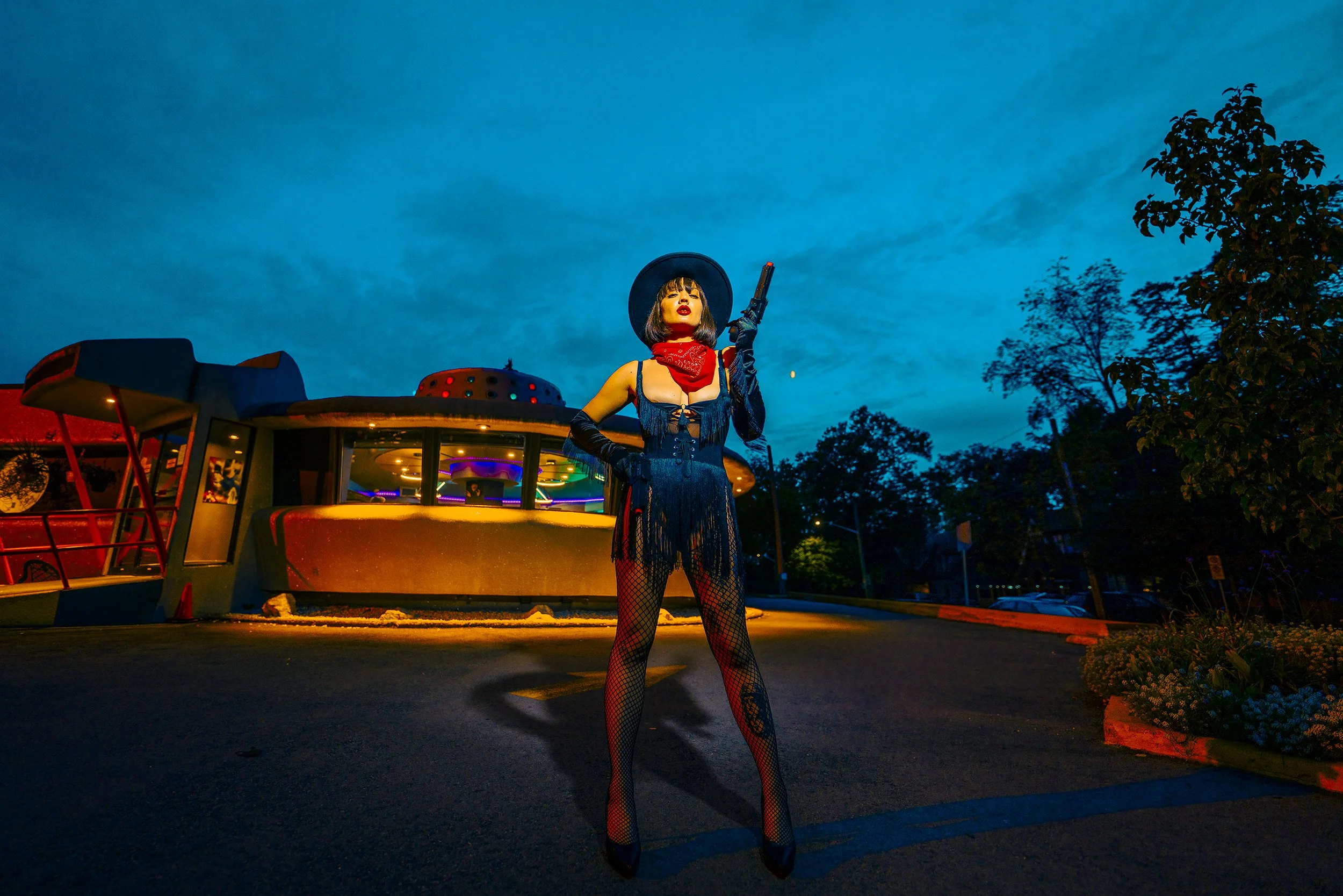A woman dressed as a cowgirl with a black hat, red bandana, and fishnet stockings stands holding a handgun, illuminated by colorful lights at night.