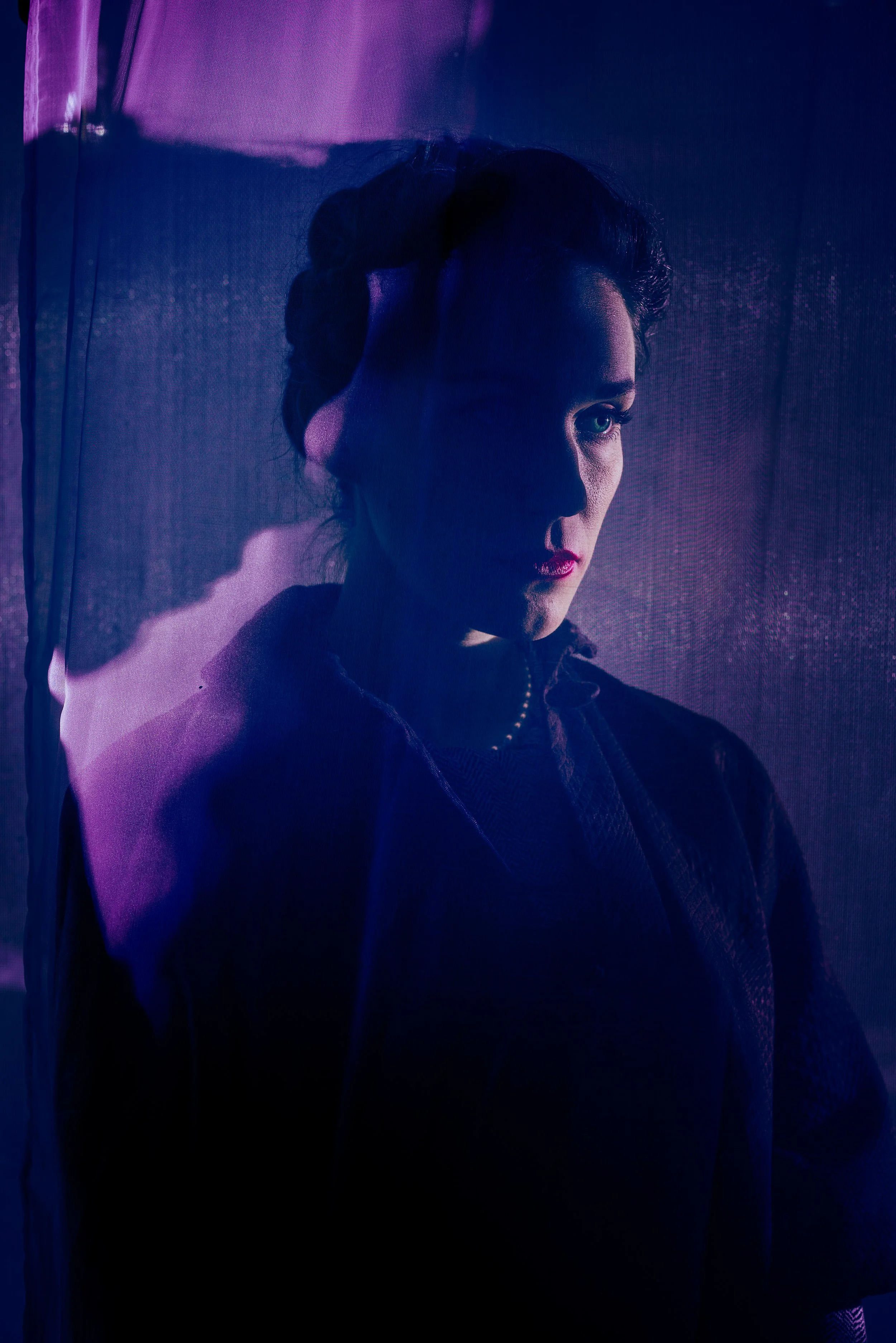 A woman with dark hair styled in an updo, wearing makeup with dark lipstick, gazes thoughtfully to the side. The image features dramatic, colorful lighting.