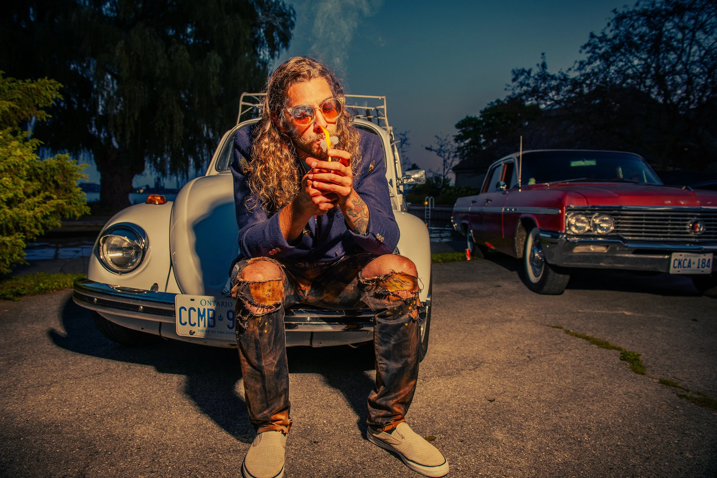 A man with long curly hair, sunglasses, and tattoos is sitting on the hood of a vintage white car with open mouth and lighting a cigarette in a parking lot during dusk. There are trees and another vintage red car in the background.