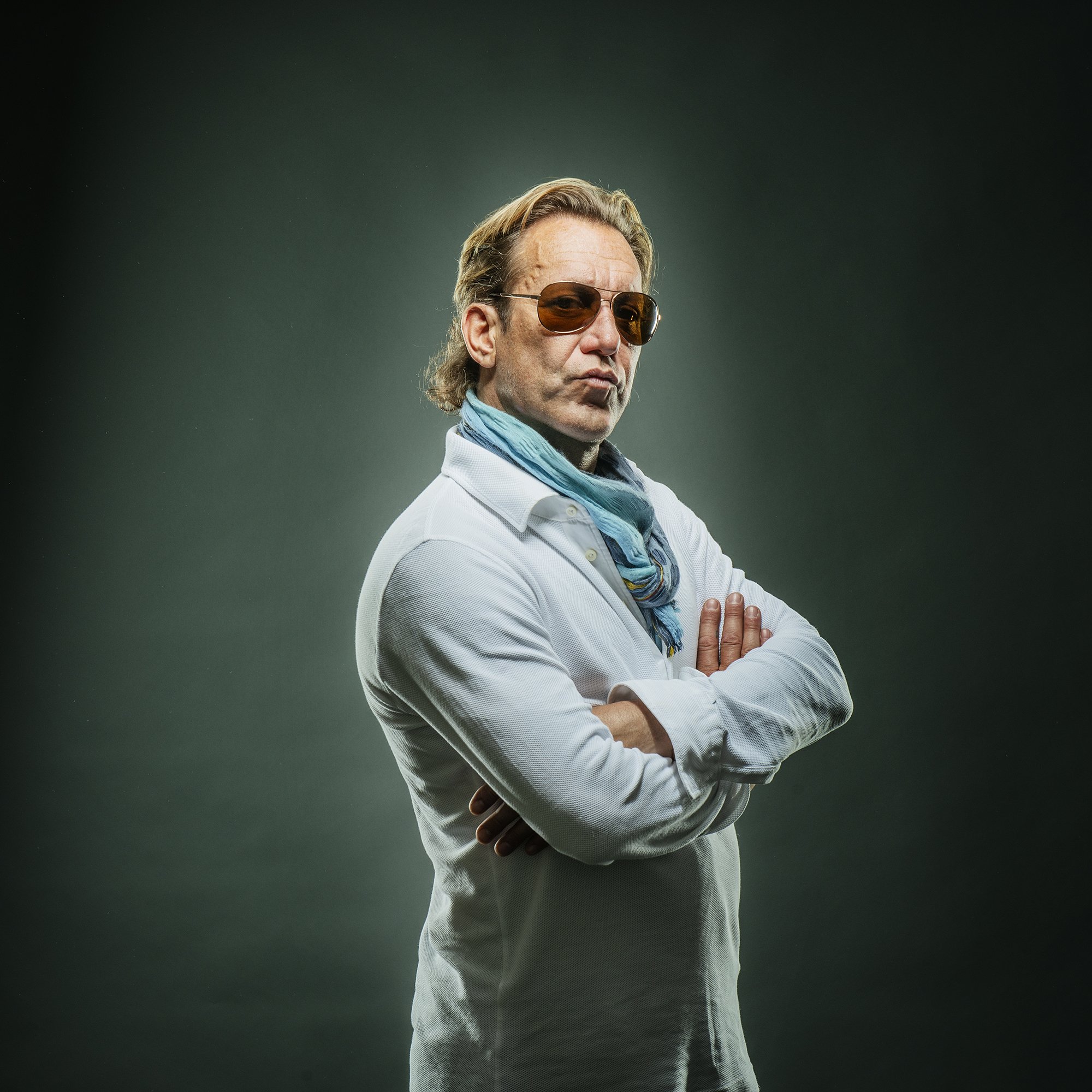 A man with light brown hair, wearing sunglasses, a light-colored shirt, a blue scarf, and crossing his arms against a dark background.