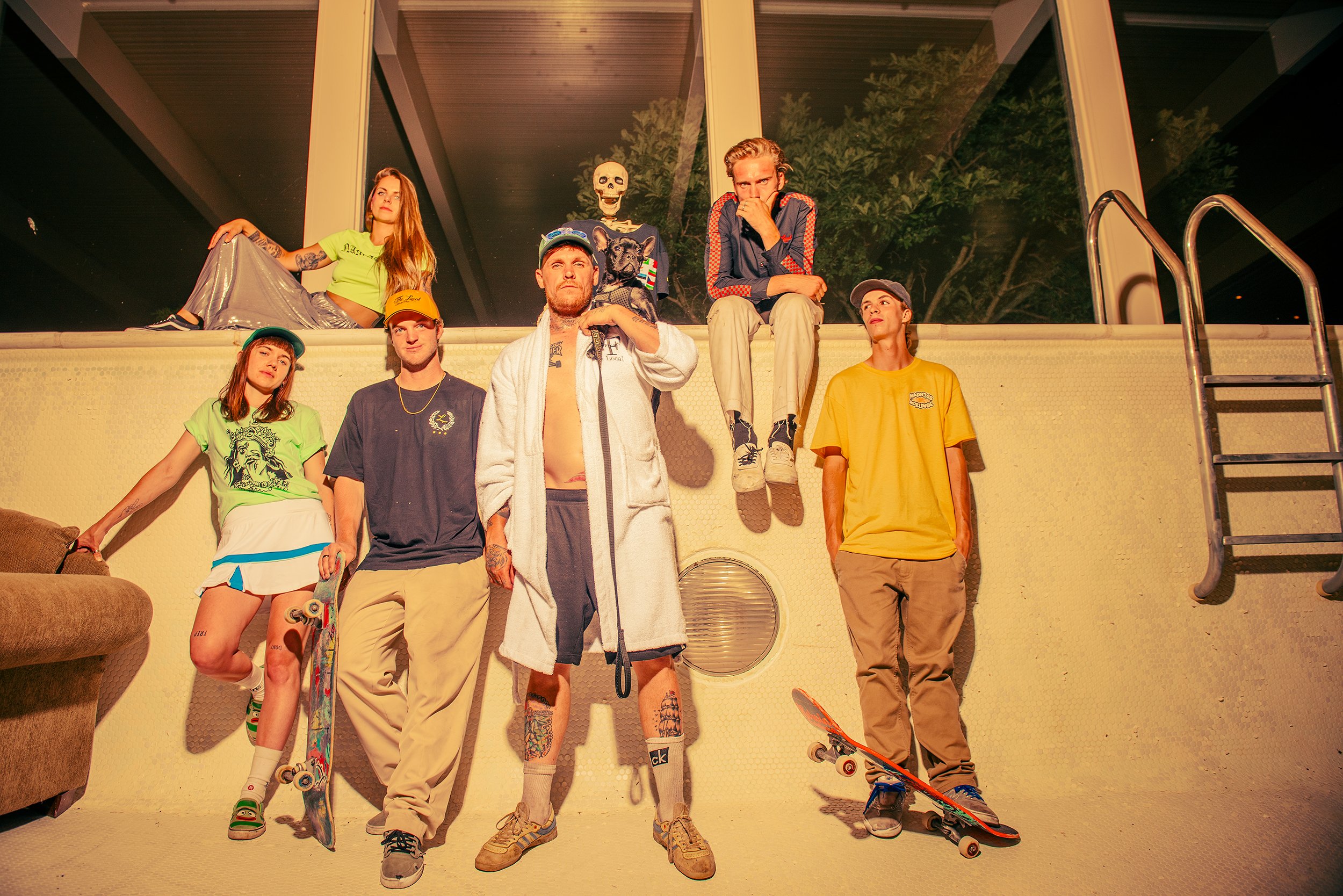 A group of nine young people, some with skateboards or skate shoes, posing on the floor and sitting on a ledge indoors near large windows, with a skeleton decoration and a dog among them.