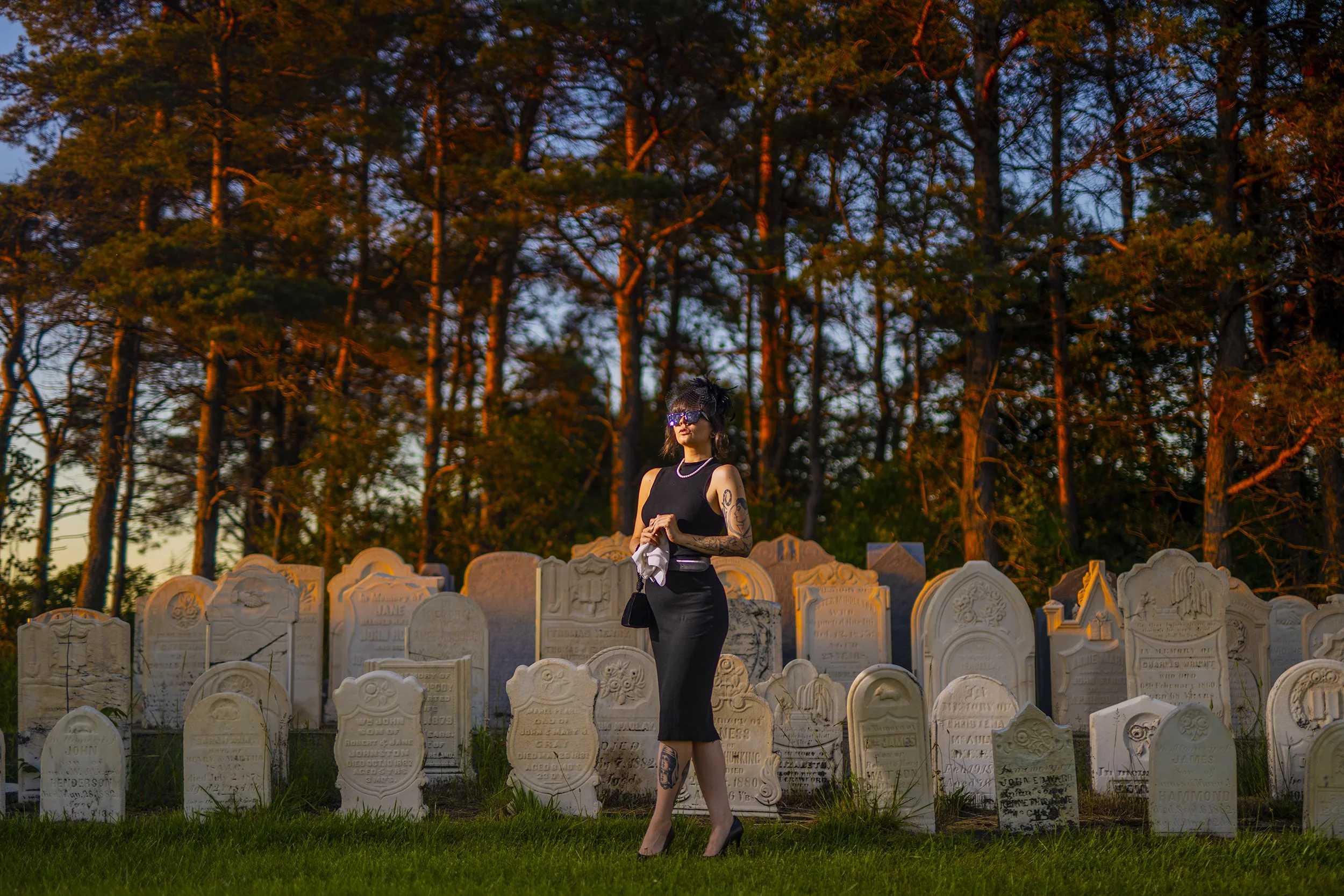 A woman dressed in black standing among tombstones in a cemetery during sunset with tall trees in the background.
