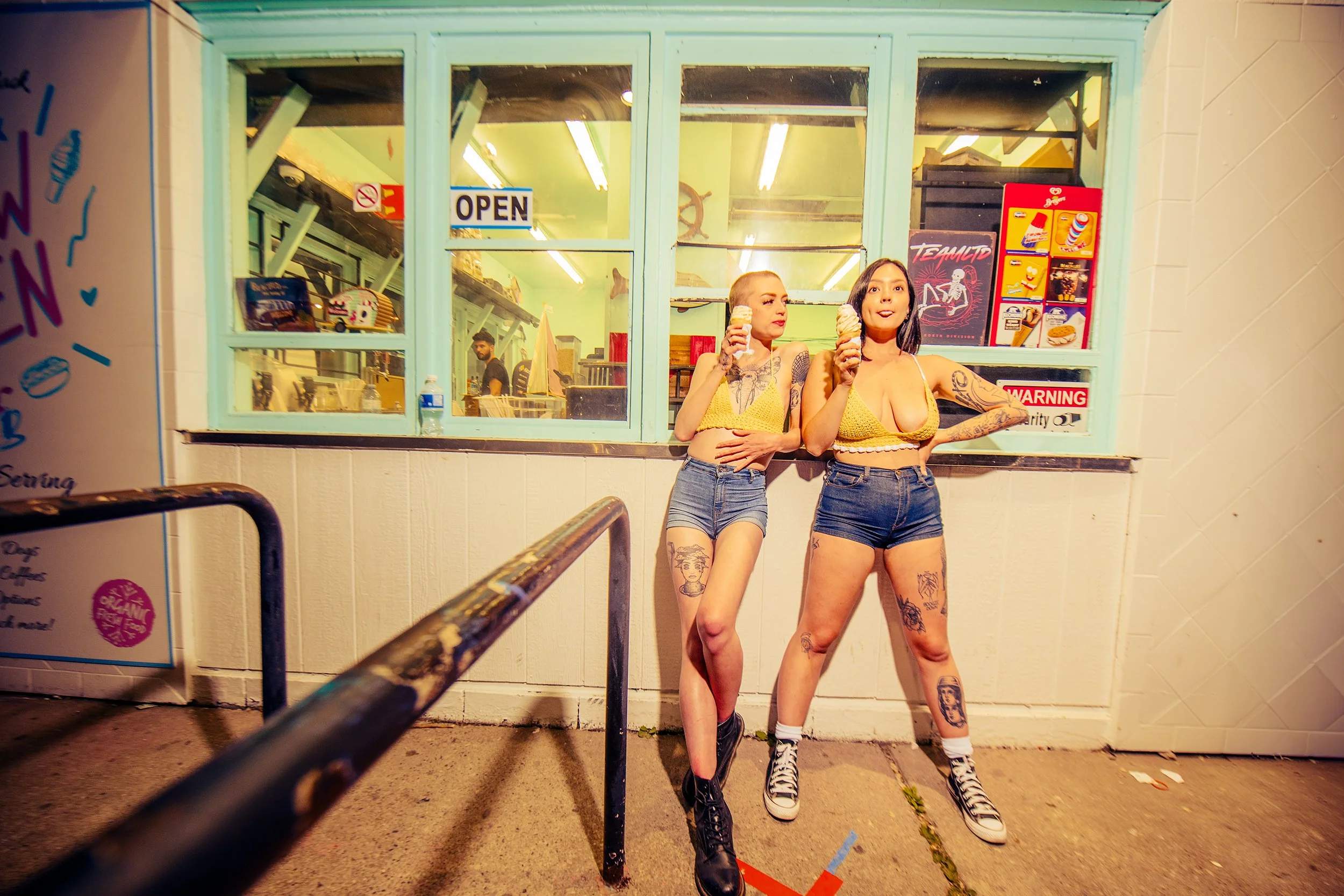 Two women with tattoos and yellow tops standing outside a colorful ice cream shop, holding ice cream cones, at night.