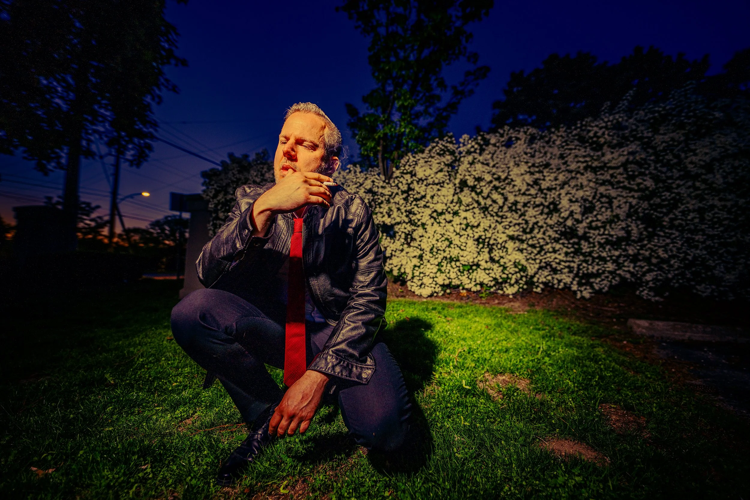 A man kneeling on the grass at night, wearing a leather jacket with a red shirt, in front of blooming white flowers and trees, with a dark blue sky and city lights in the background.