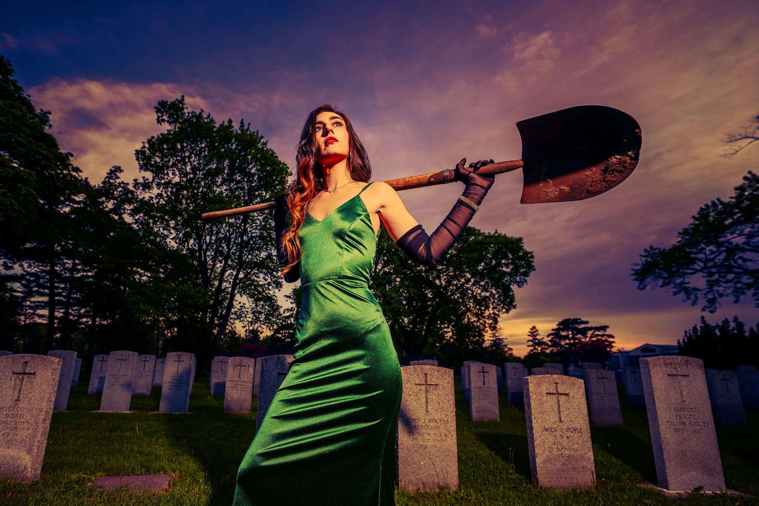 A woman in a green satin dress holding a shovel over her shoulder, standing in a graveyard at sunset with tombstones behind her and trees in the background.