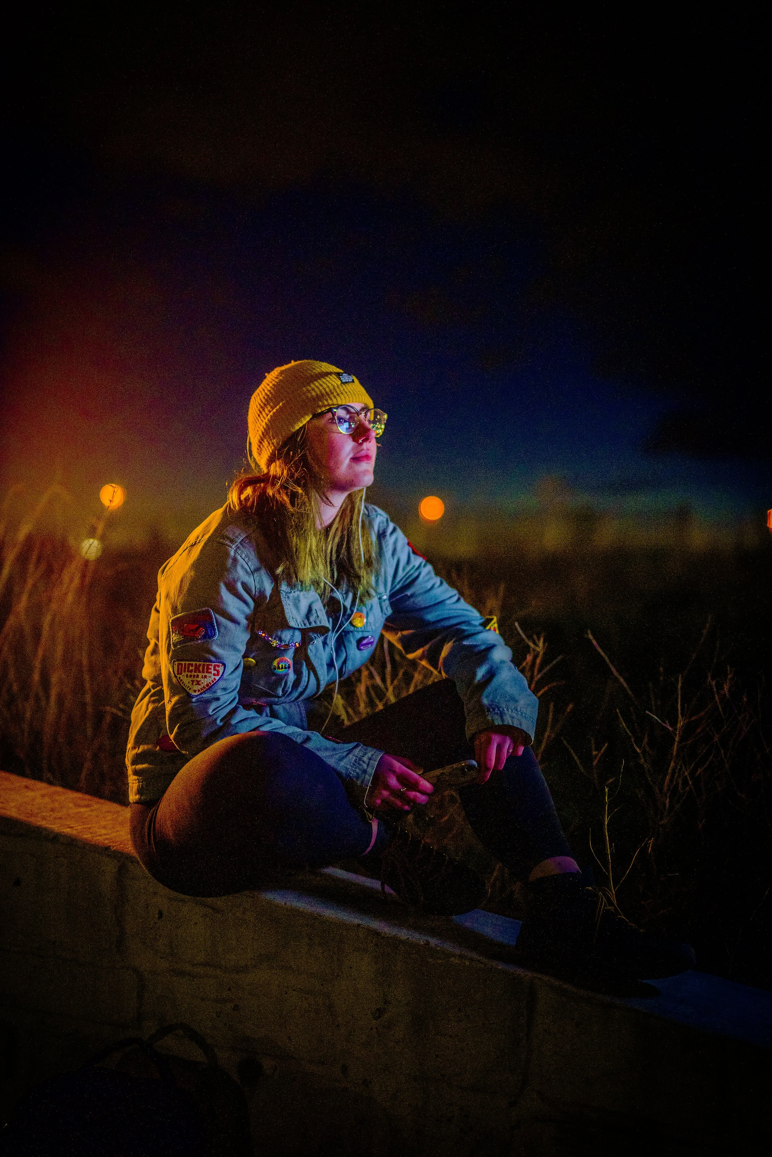 A woman with glasses and a yellow beanie sitting outdoors at night, illuminated by warm light, with a serious expression, surrounded by tall grass, under a dark starry sky.