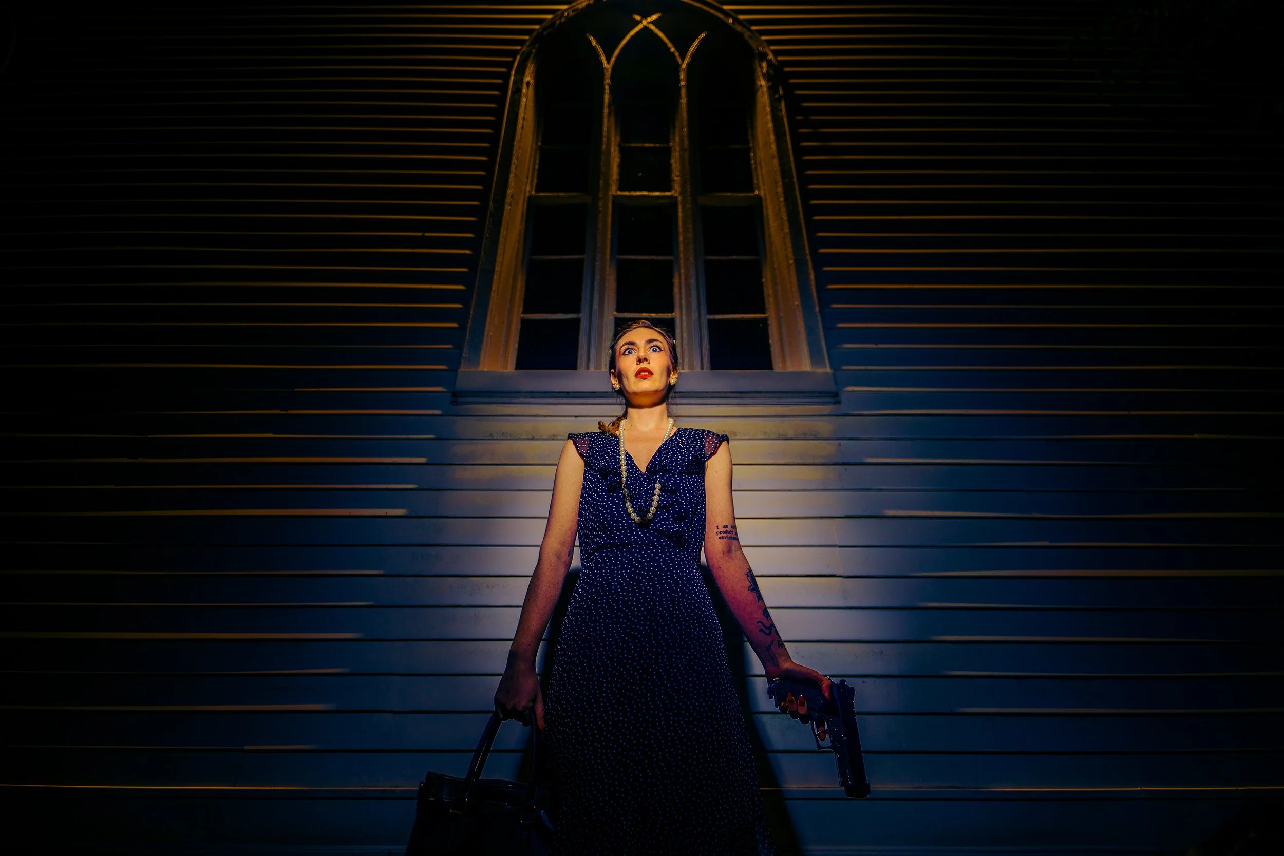 A woman with dark hair and bright red lipstick standing outdoors at night in front of a wooden wall with a large arched window. She is wearing a sleeveless navy polka dot dress, pearl necklace, and has visible tattoos on her arms. She is holding a bl