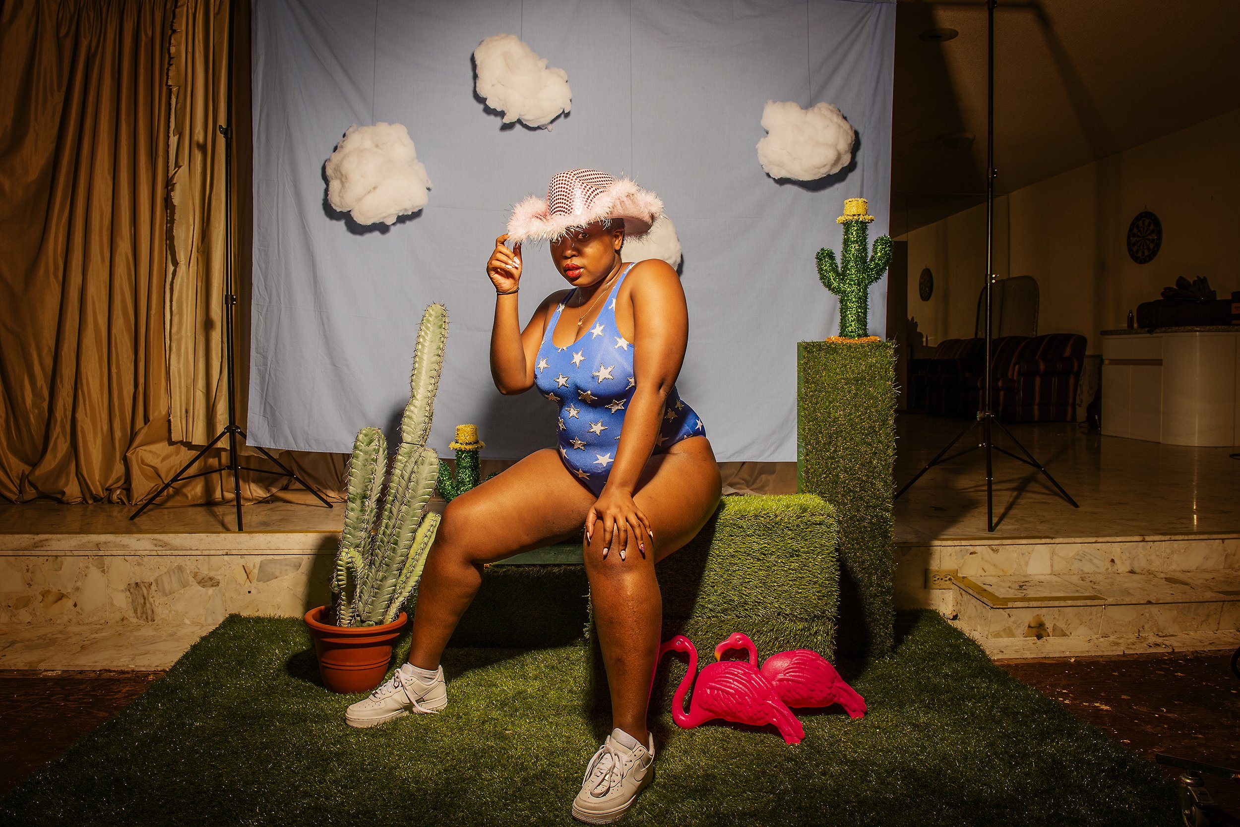 A woman wearing a star-spangled bathing suit and a pink cowboy hat, sitting on a grassy platform with pink flamingo lawn ornaments, surrounded by cactus decorations, in front of a backdrop with cotton cloud props.
