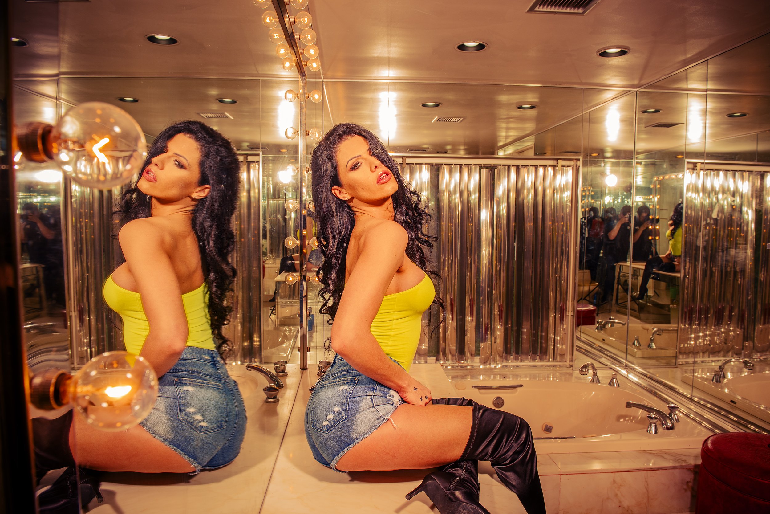 A woman with long dark hair taking a mirror selfie in a dressing room, wearing a yellow top, denim shorts, and black boots, with a background of mirrors and warm lighting.