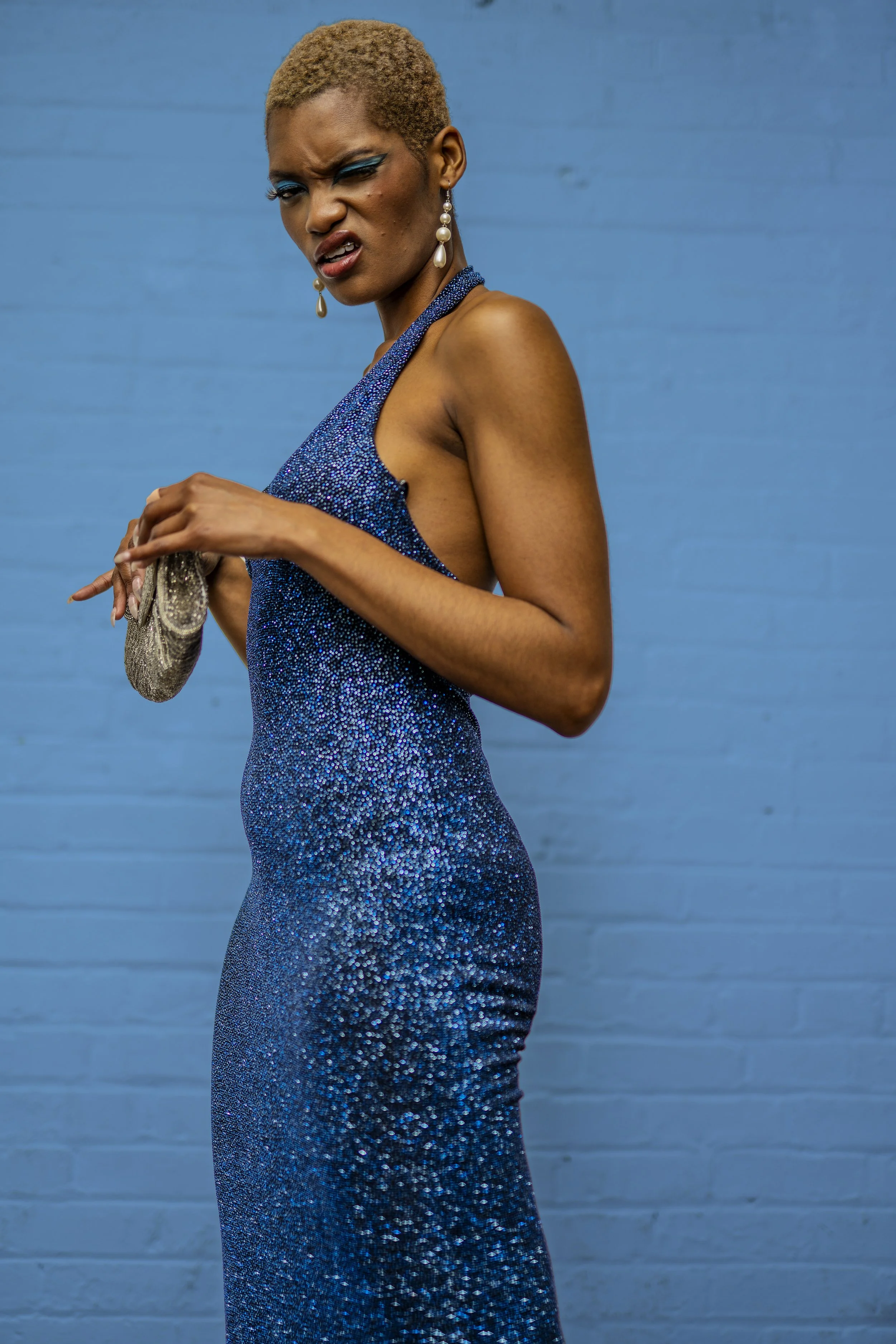 A woman with short blonde hair wearing a form-fitting, sparkling blue dress and pearl earrings, standing against a blue wall and making a face.