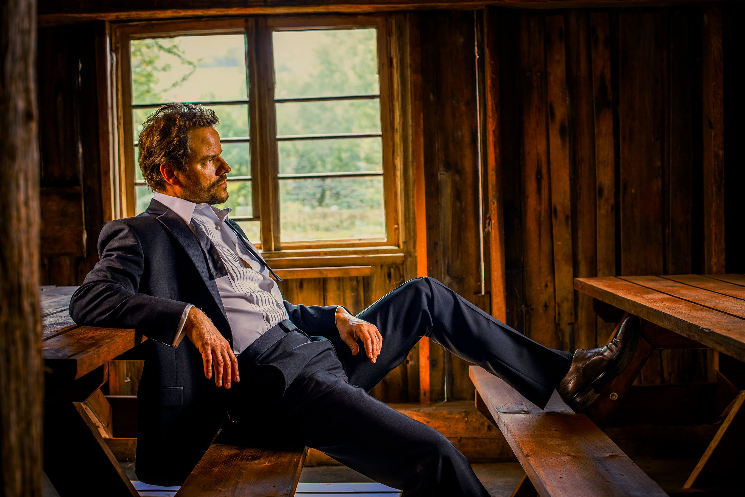A man in a business suit sitting relaxed inside a rustic wooden cabin, with a large window showing greenery outside.
