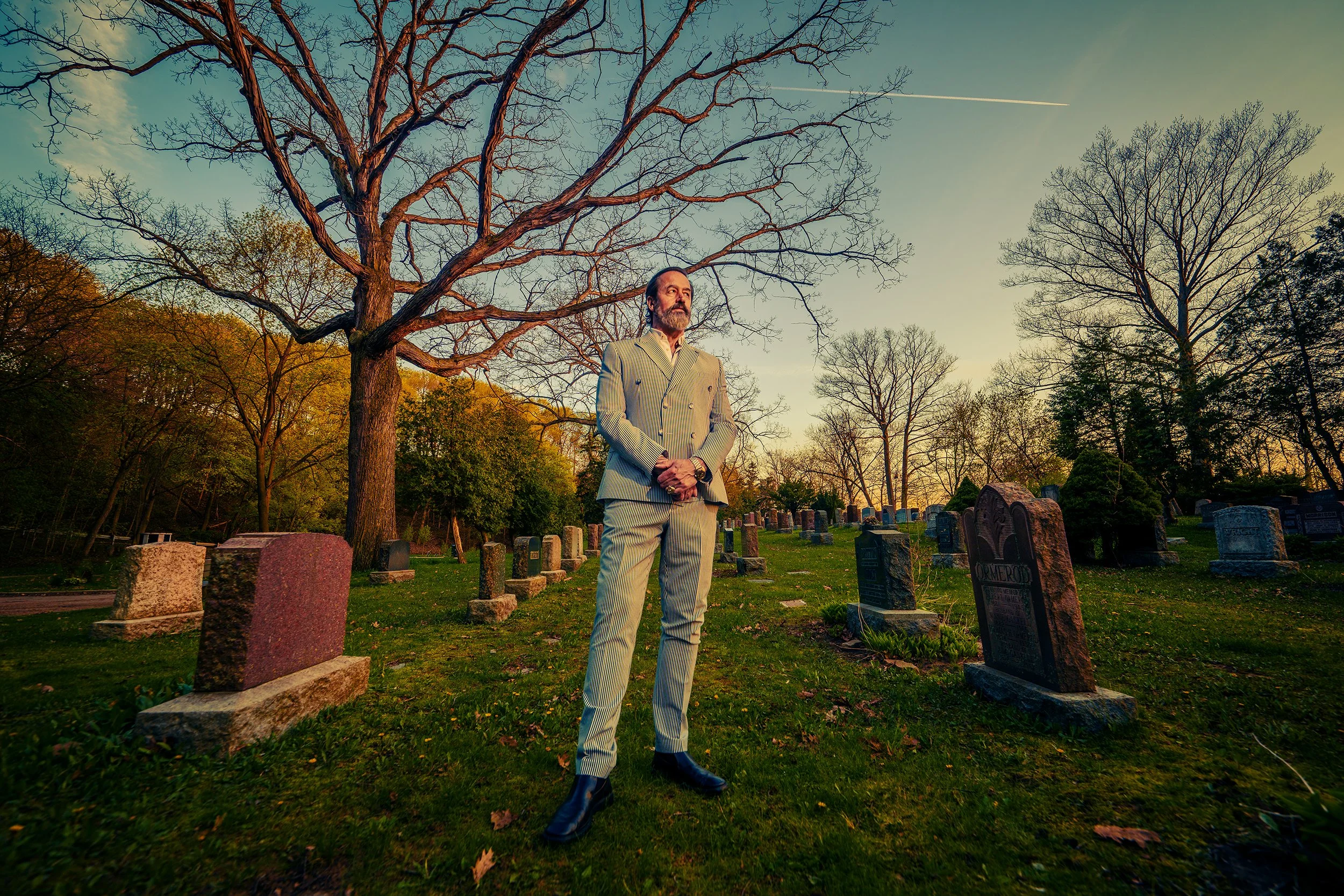 A man in a striped suit standing in a cemetery at sunset with trees in the background and gravestones around him.