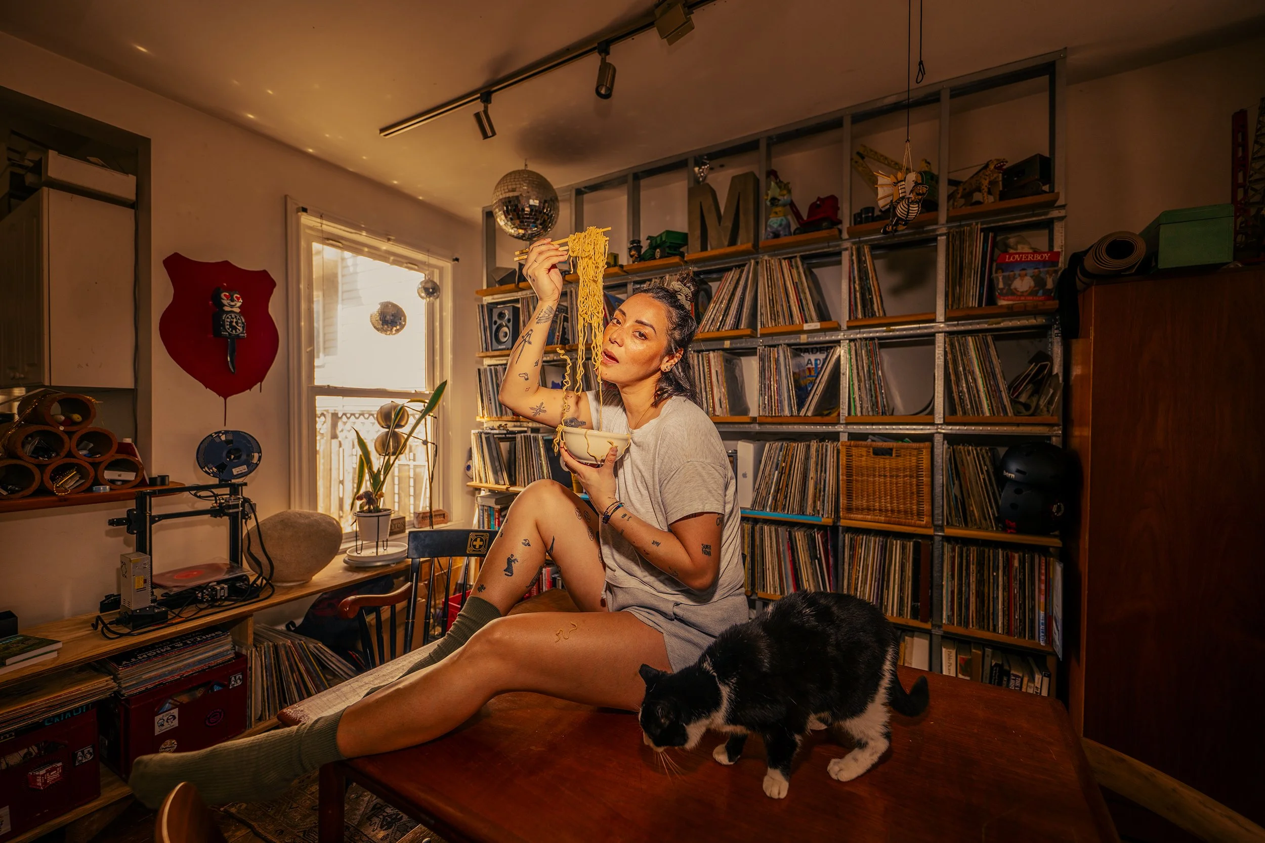 A woman sitting on a table in a cozy, music-themed room, holding a bowl of noodles with chopsticks, with a black and white cat nearby and a large collection of vinyl records behind her.