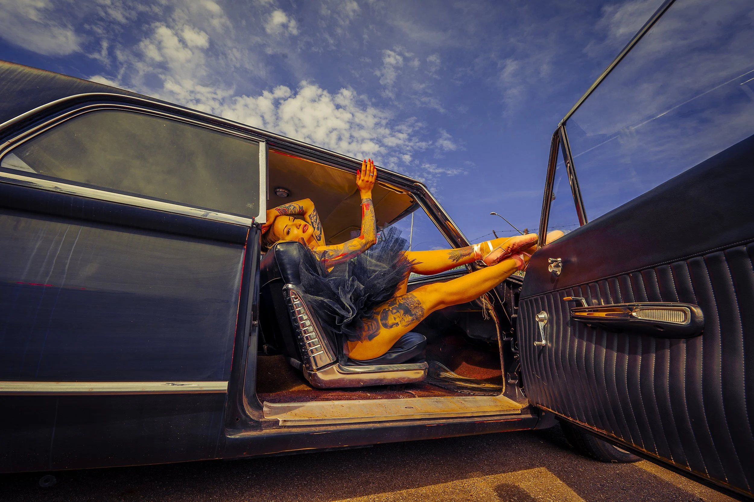 A woman with tattoos in a black dress and high heels lying in the back seat of a vintage black car with the door open, under a blue sky with scattered clouds.