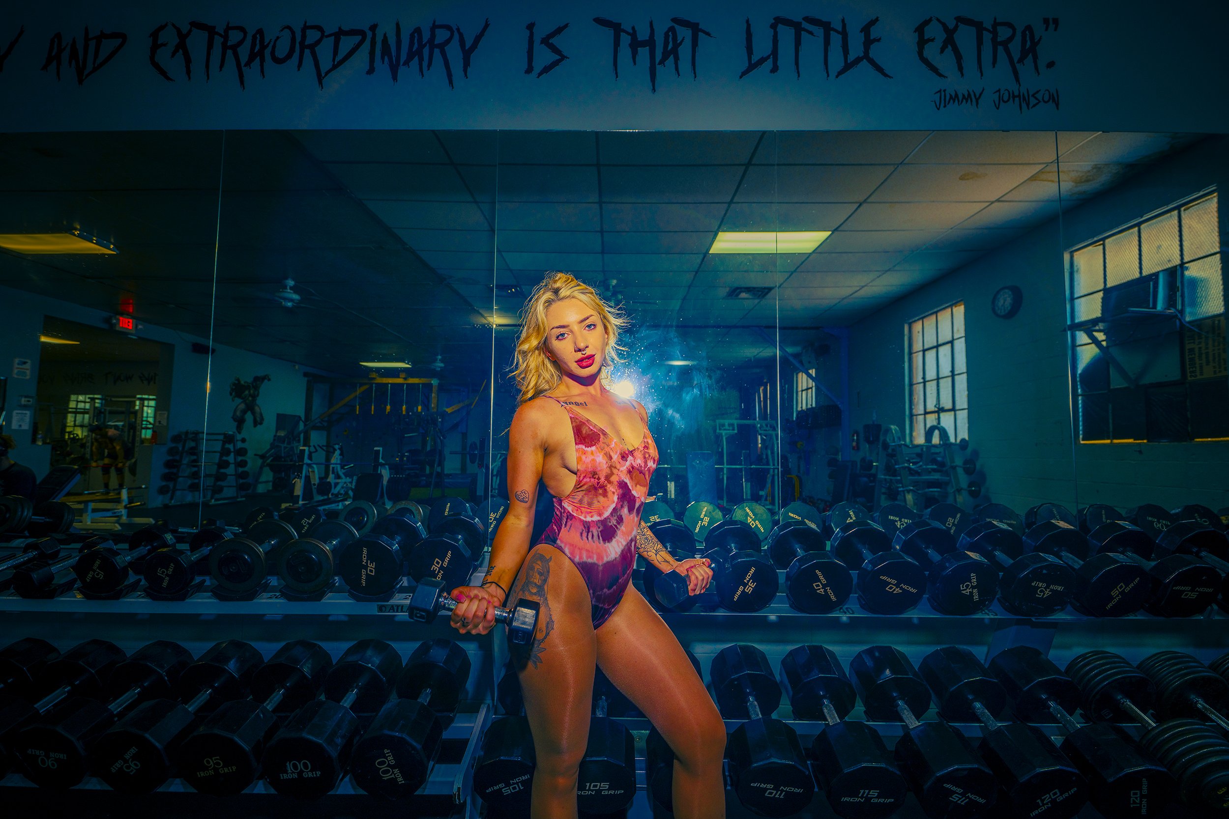 A woman with tattoos on her arms and legs is lifting dumbbells in a gym. She is wearing a colorful, patterned one-piece swimsuit. The gym has a mirrored wall behind her, with rows of dumbbells on racks, and bright windows on the side. A quote by Jimm