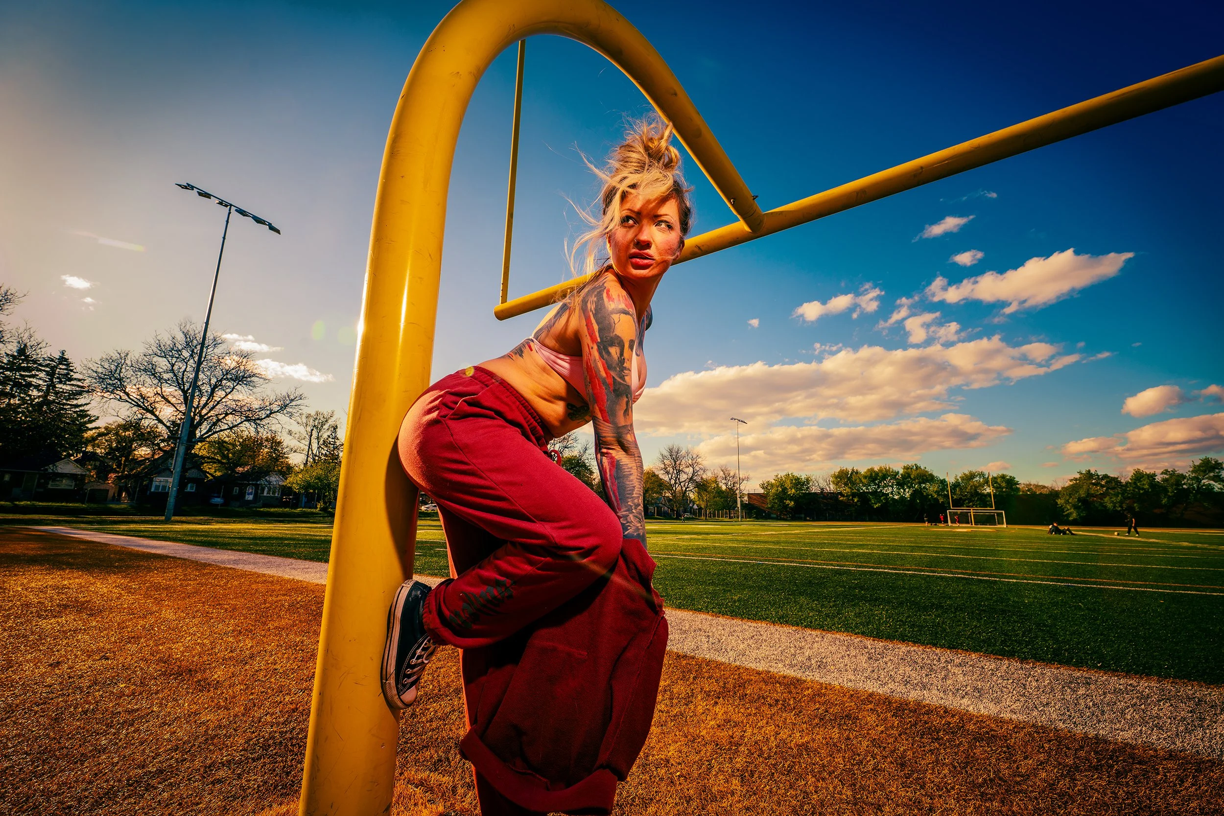 A woman with tattoos is kneeling on the ground at a sports field, leaning against a yellow goalpost, during sunset with a partly cloudy sky.