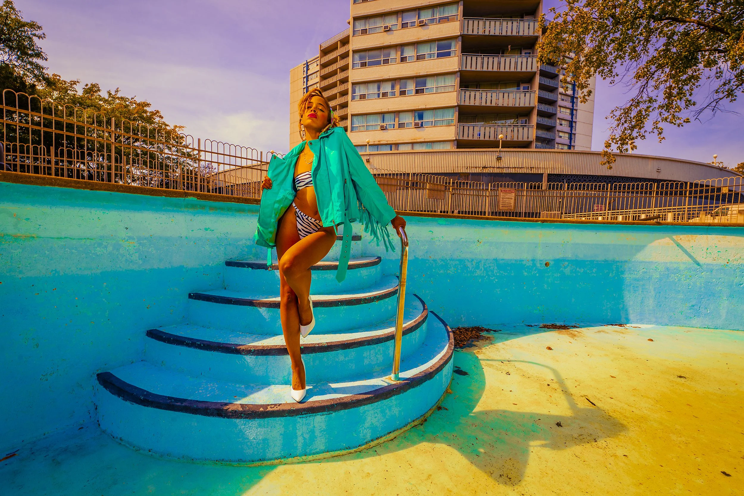 A woman in a zebra print swimsuit and white high heels poses on the steps of an empty swimming pool, wearing a teal jacket draped over her shoulders. The pool is painted in bright colors and the background features a modern apartment building, trees,
